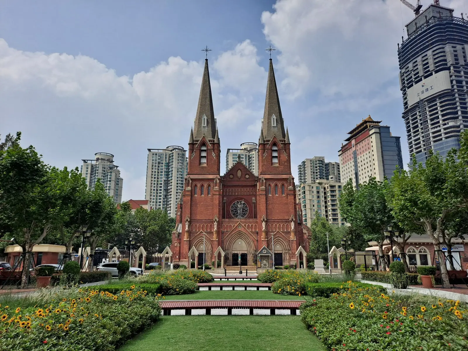 A red-brick Gothic cathedral with two tall spires and a rose window, seen across a flower garden with high-rise buildings behind.