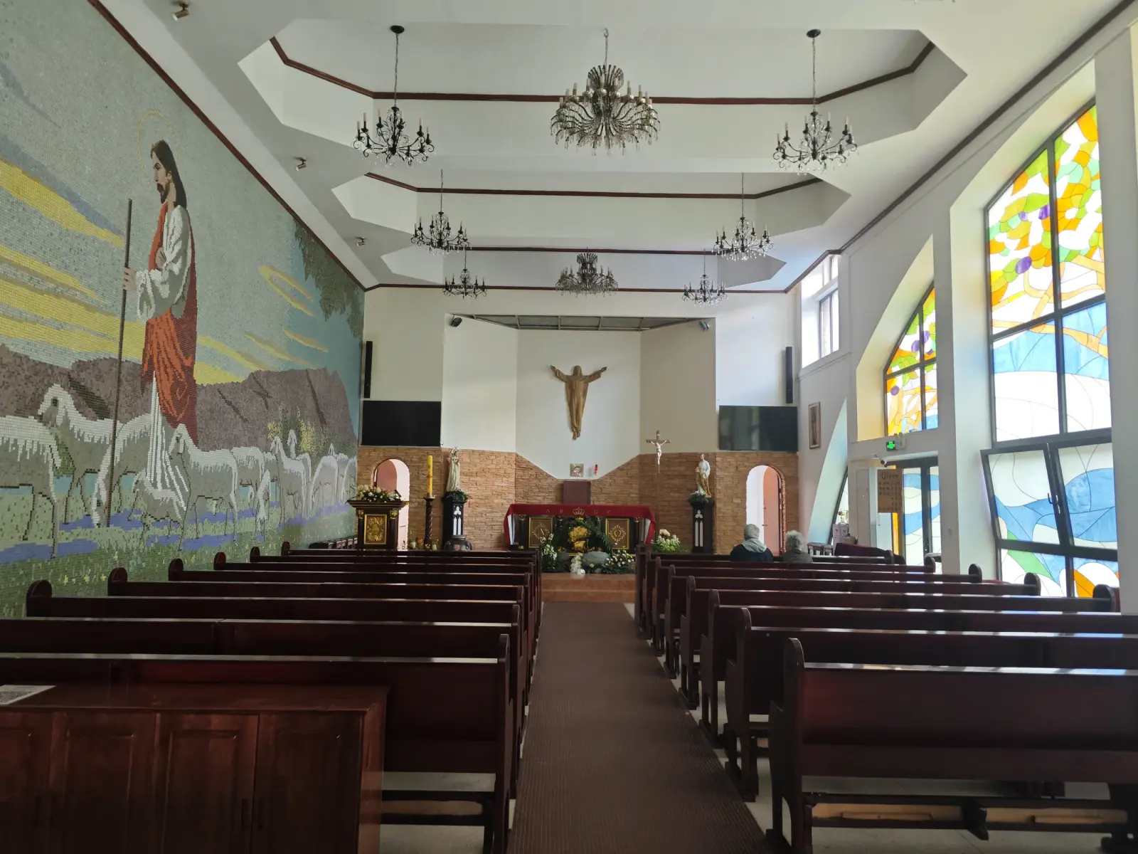 Church interior with wooden pews, a crucifix over the altar, stained glass and a mosaic wall.