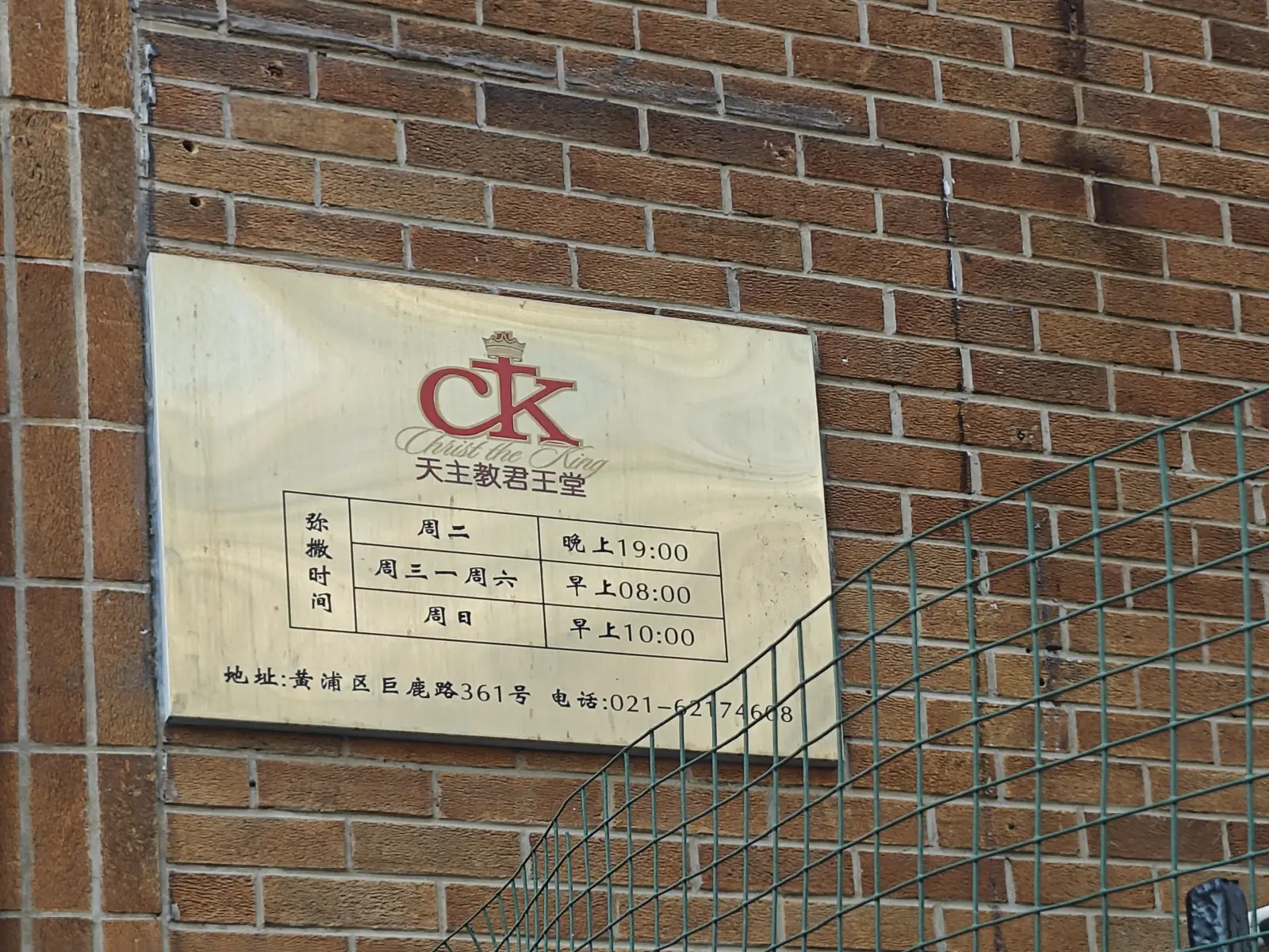 A brass-coloured plaque on a brick wall listing Mass times for Christ the King Church.