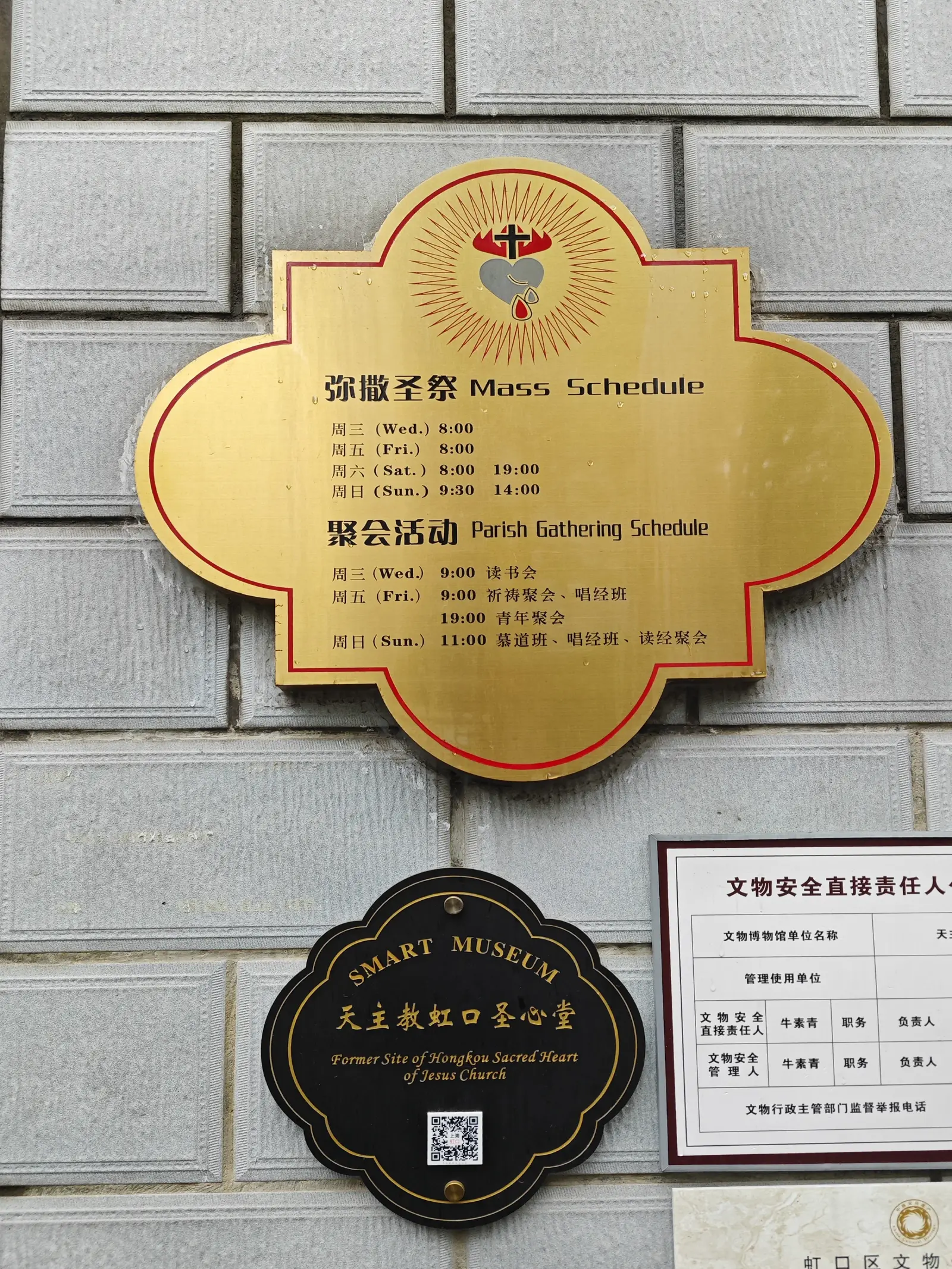 A gold plate with Mass times in Chinese and English above a black plaque on a grey brick wall.