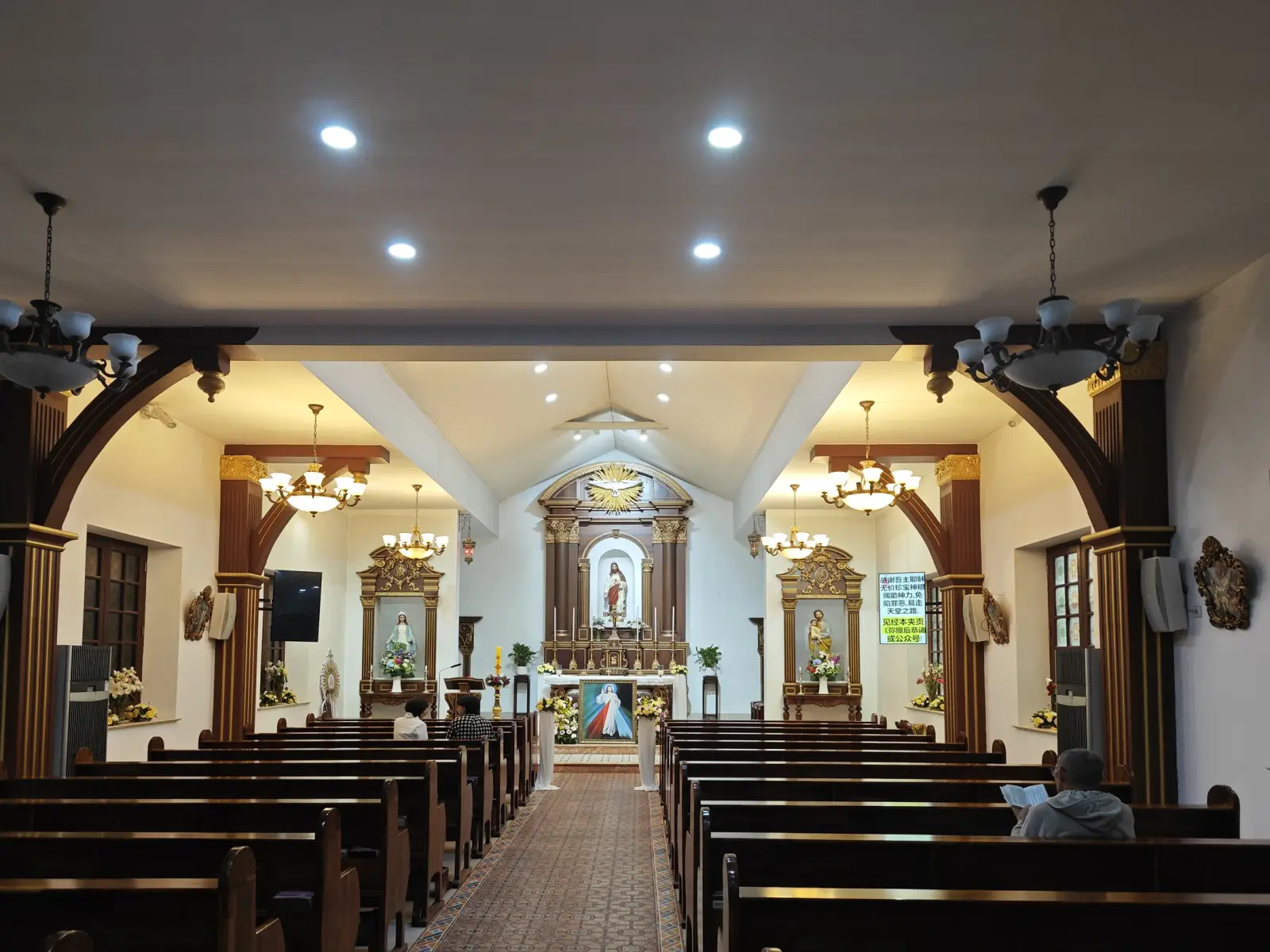 A church interior with wooden pews, chandeliers and a white altar with a statue of the Sacred Heart.