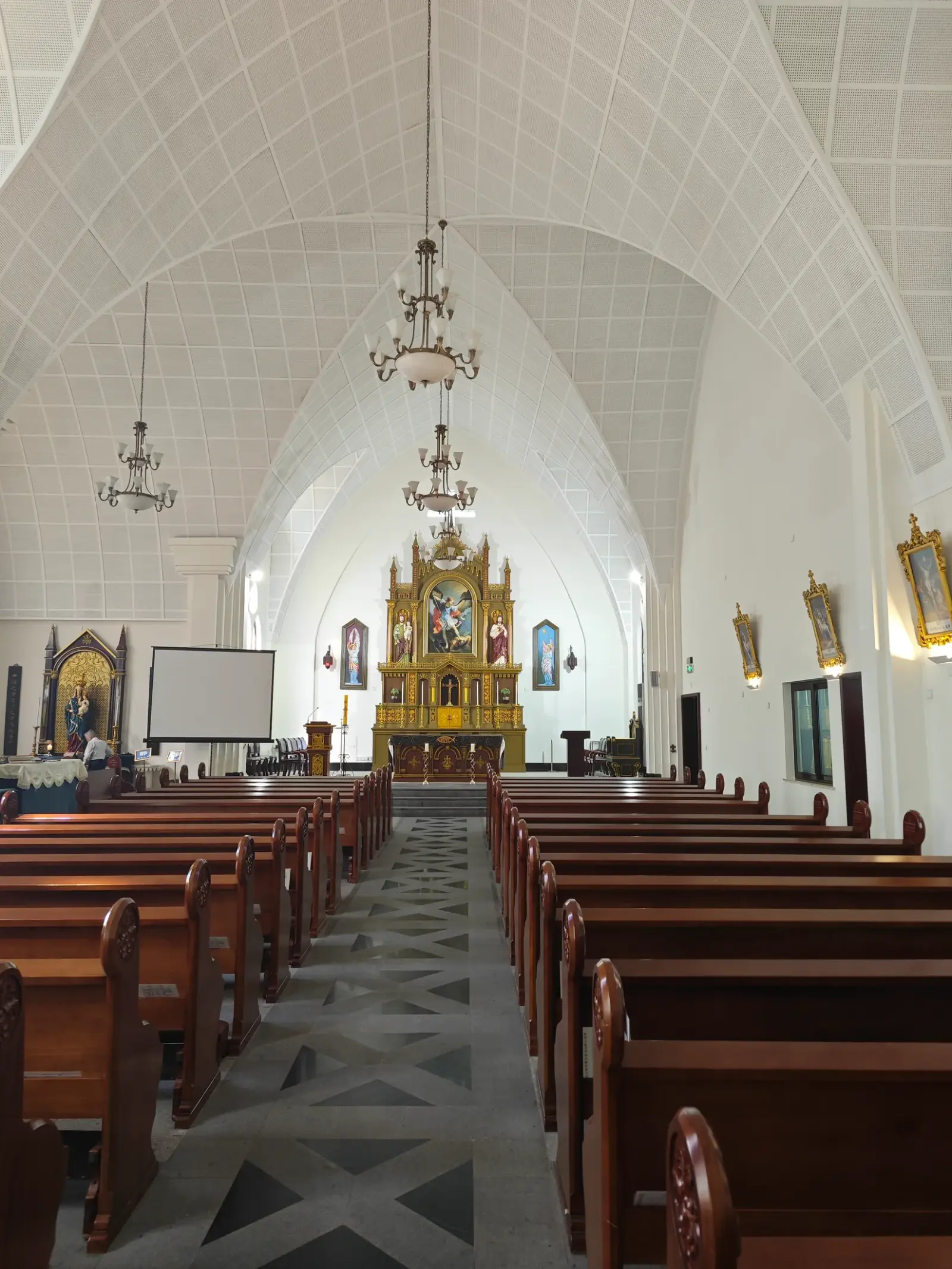 A church interior with a white ribbed vault, wooden pews, chandeliers and a golden altar.