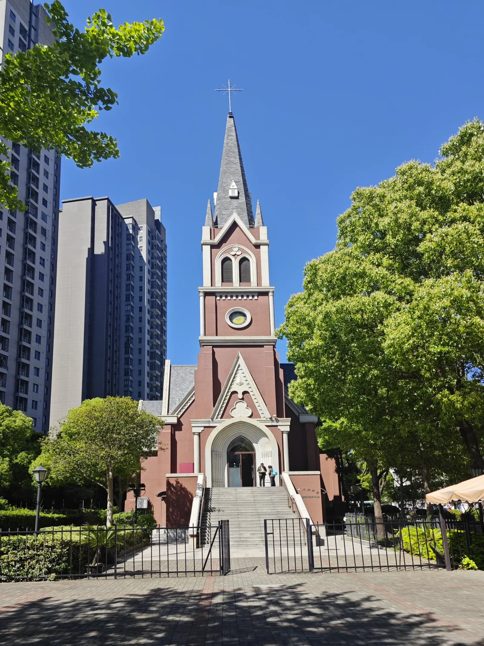 A red-brick Gothic-style church with a tall slender spire, a clock and a pointed doorway, with blue sky and trees around it.
