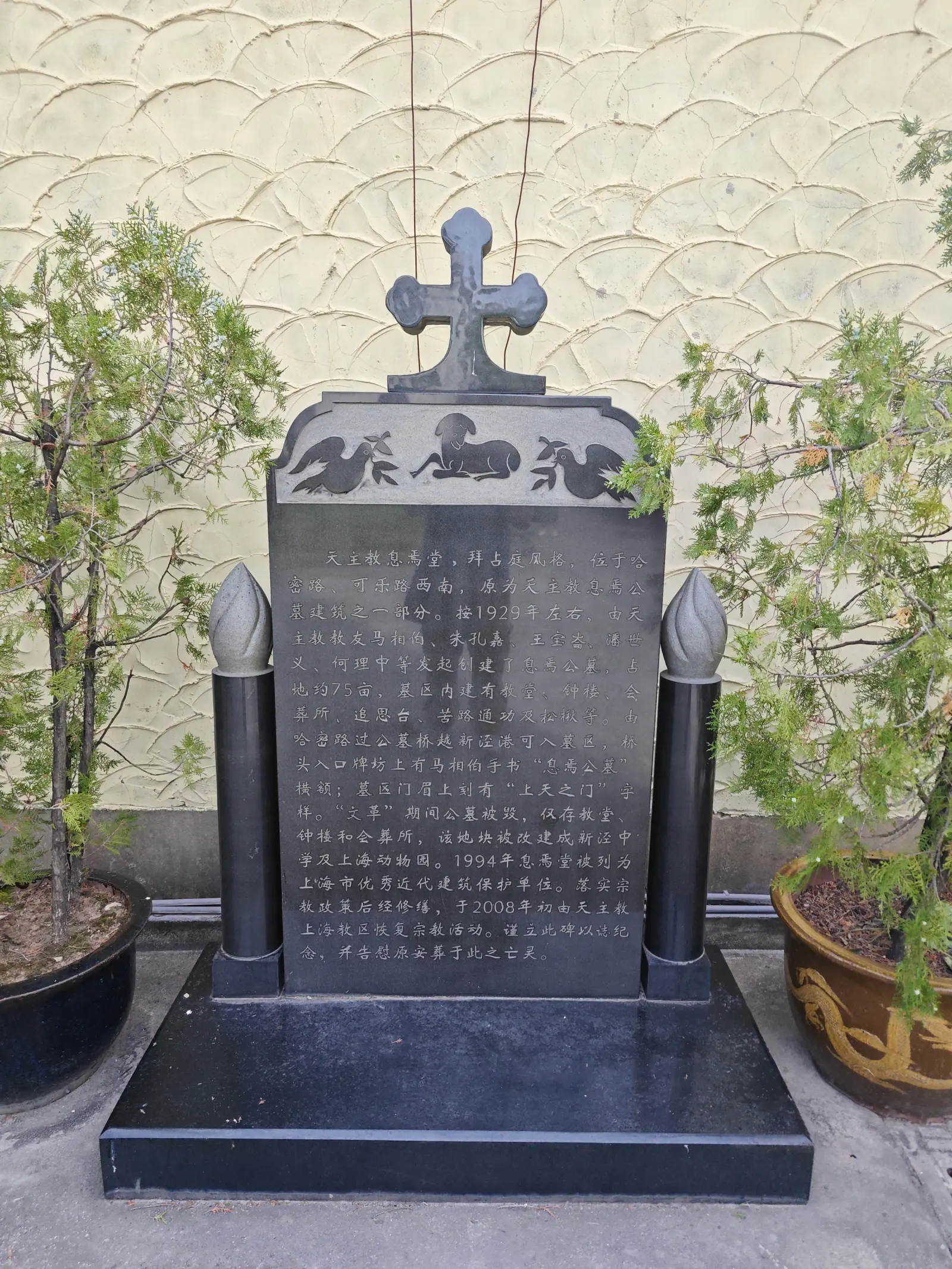 A black memorial tablet topped by a cross between two lamp-like columns, against a scalloped wall.