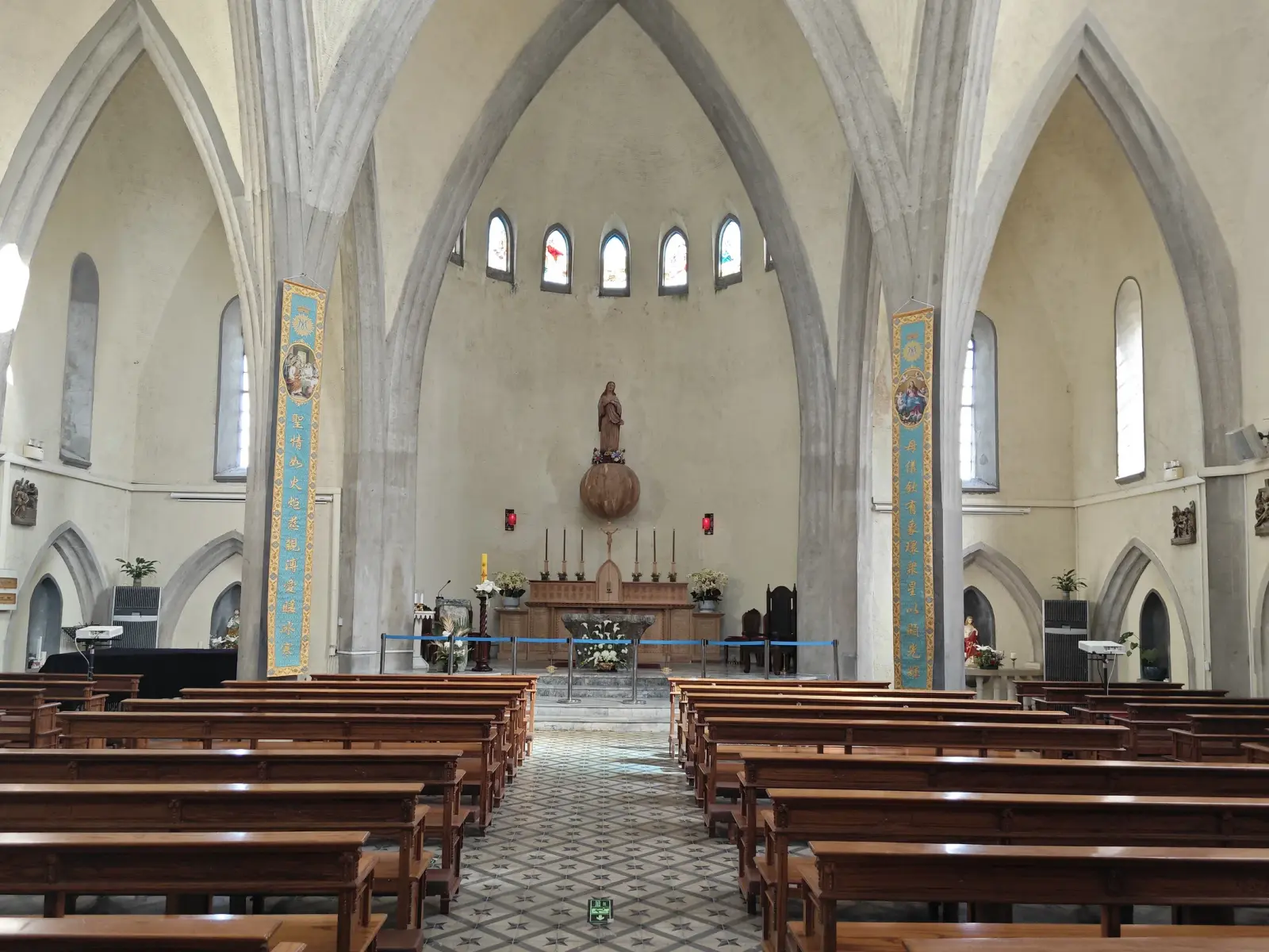 A pale church interior with large pointed arches, wooden pews, a tiled aisle and a statue at the end.