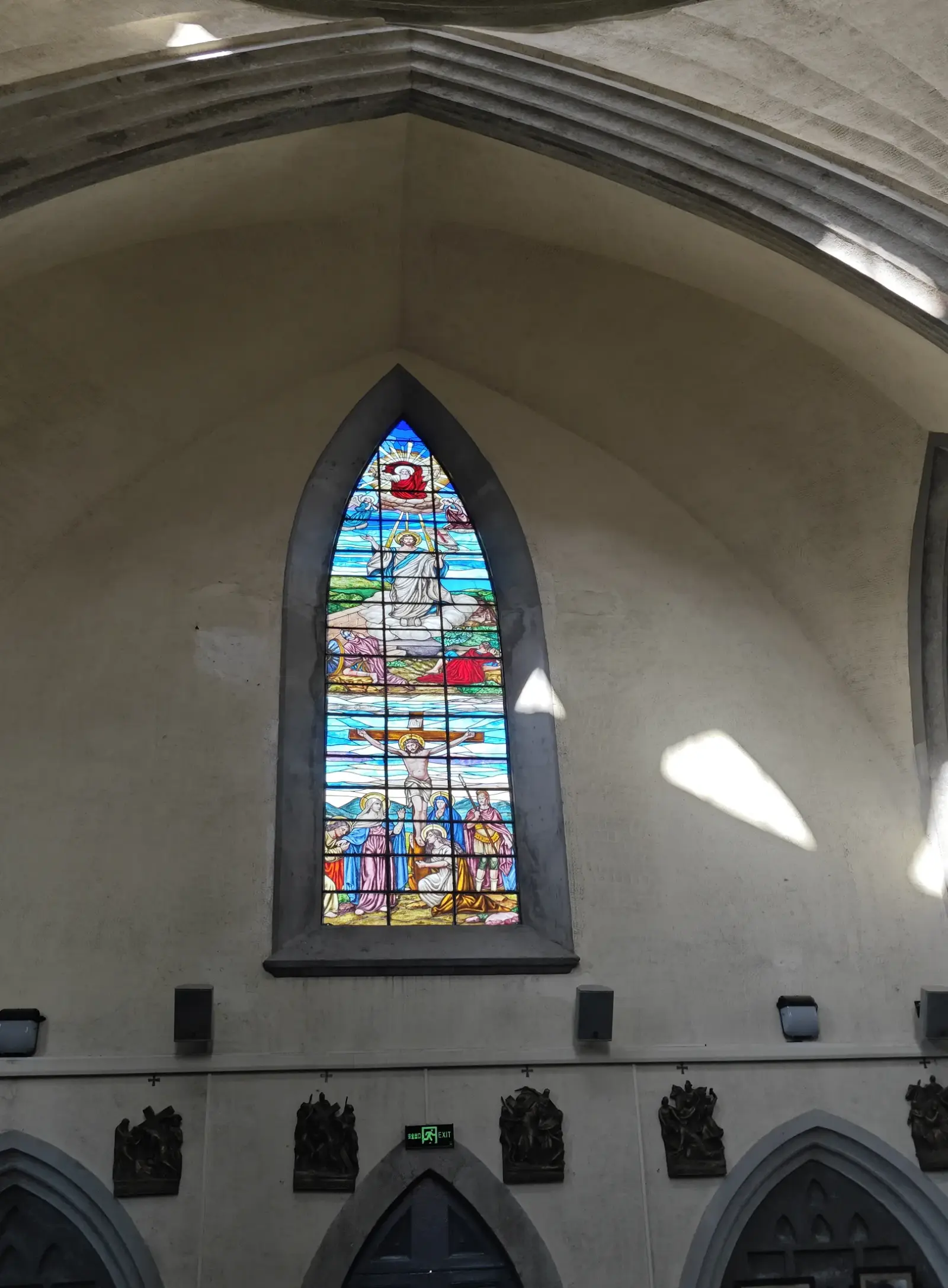 A narrow stained-glass window in a pale interior wall above small dark relief panels.