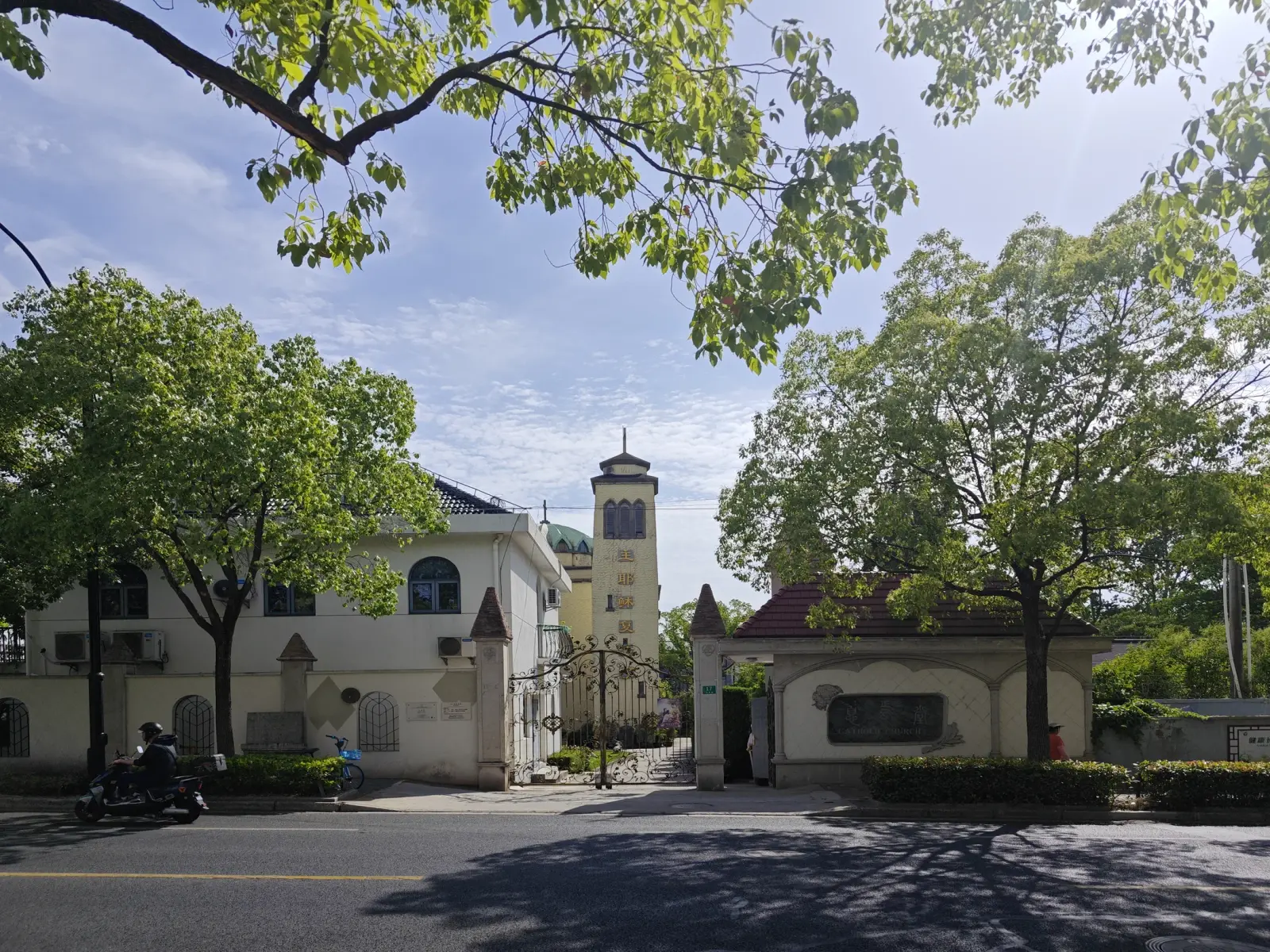 A tree-lined road with a cream compound wall, an iron gate and a church tower behind.
