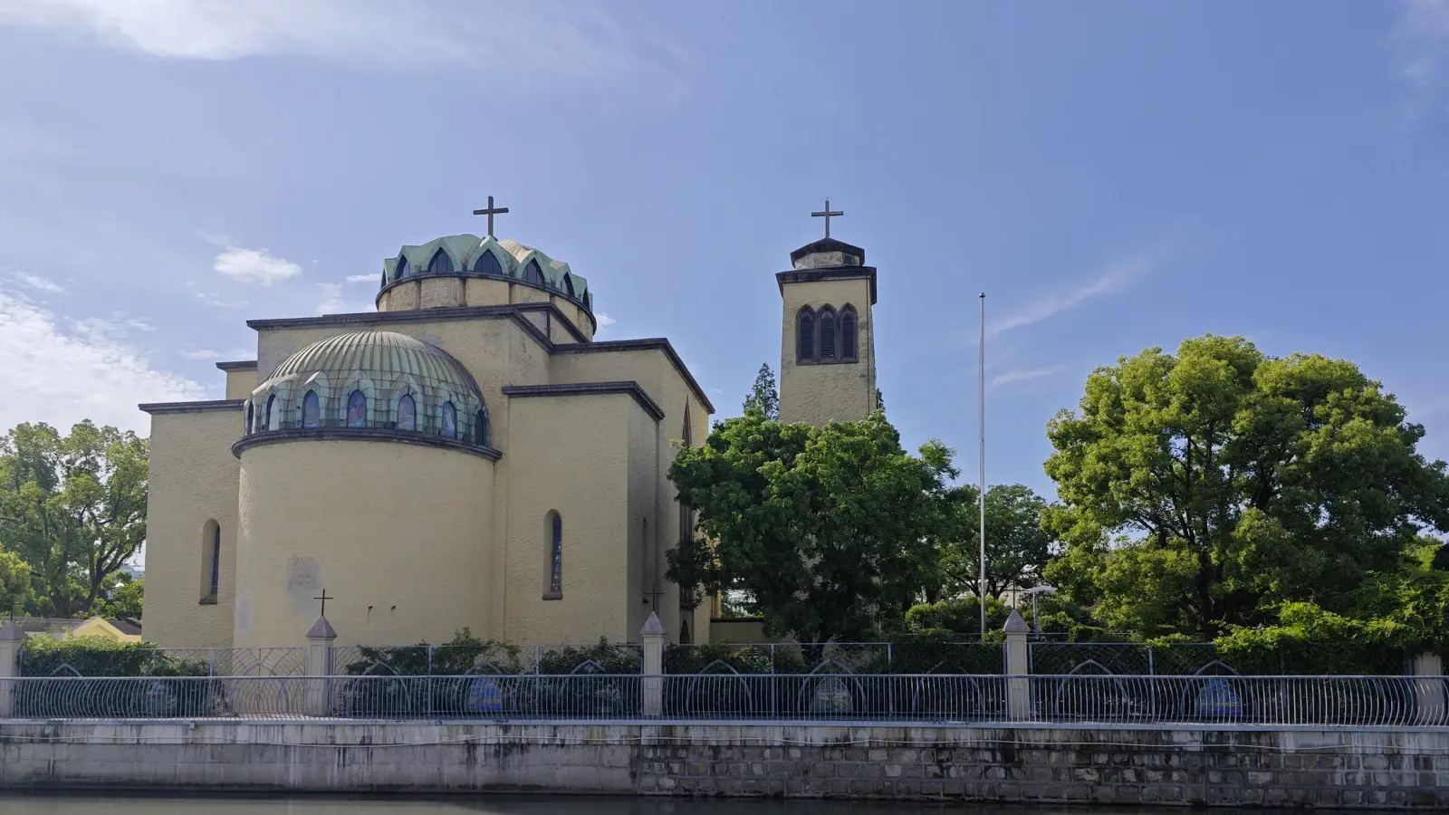 A yellow-ochre church with two green domed roofs and a square tower, behind trees and a canal-side fence under a blue sky.