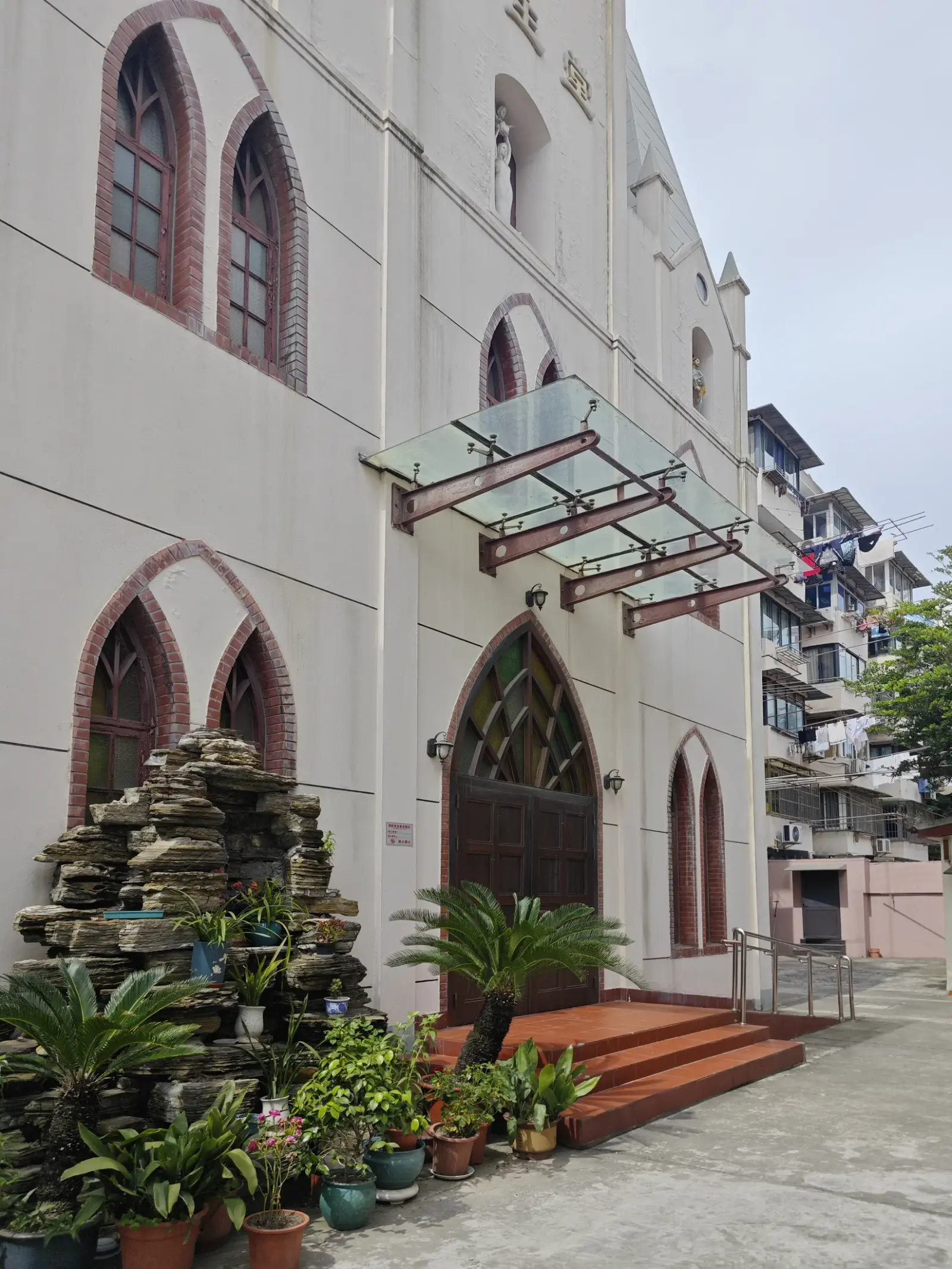 A church entrance with a pointed arch door, glass canopy and potted plants beside a rock garden.