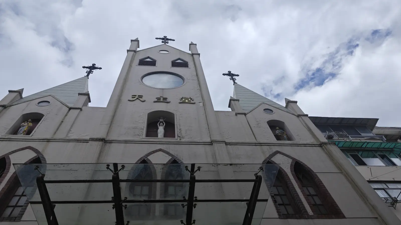 The upper facade of a white church with a round window, statue niches and three crosses against clouds.