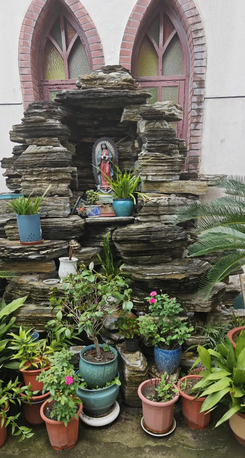 A rock grotto with a small blue-robed statue among potted plants in front of arched windows.