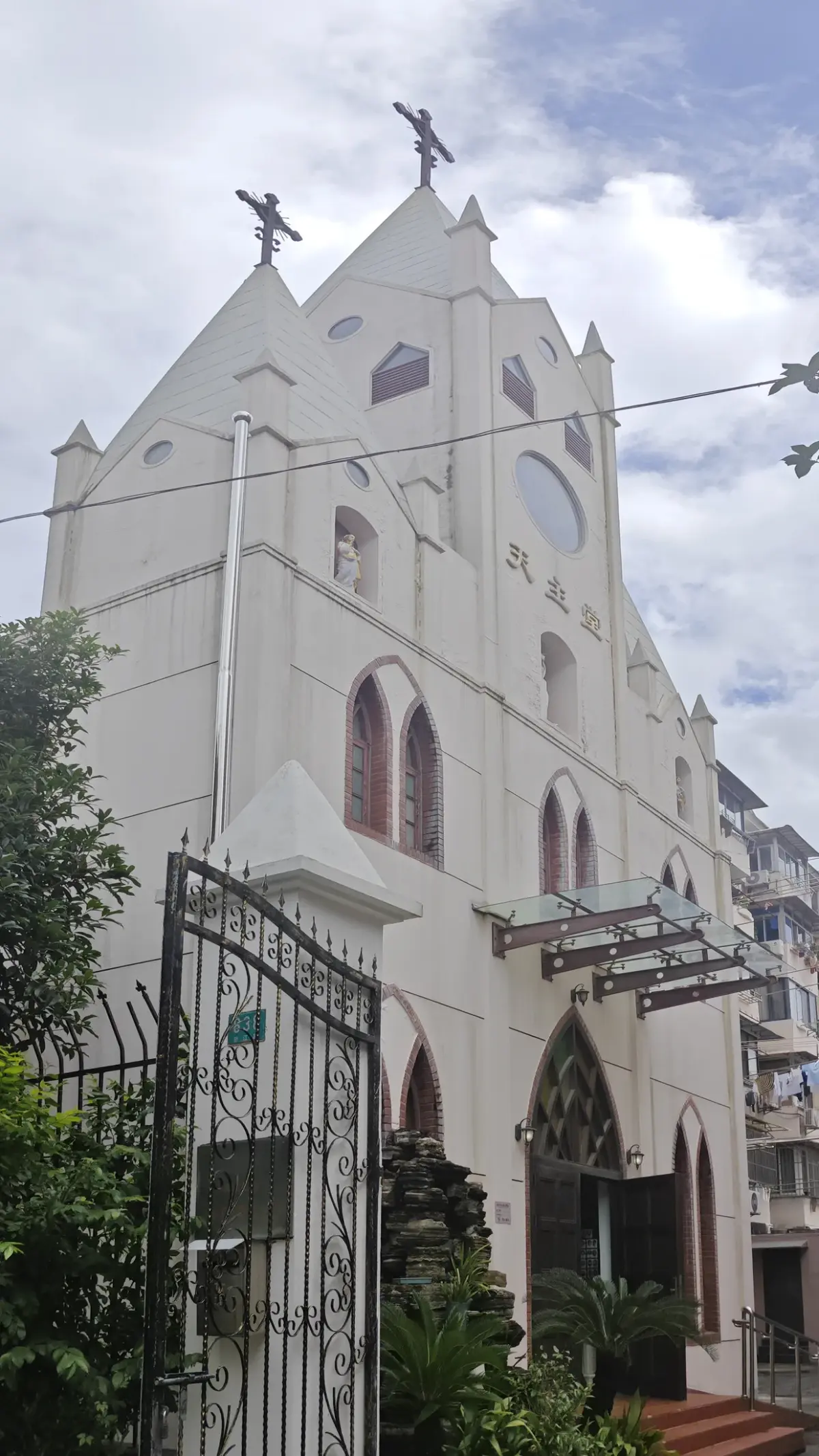 A white church facade with pointed gable, round window, pointed arched windows and crosses, seen from beside a wrought-iron gate.
