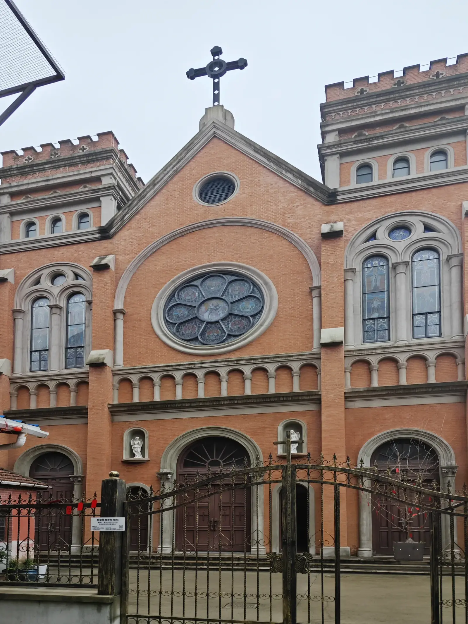 An orange-brick church facade with two squat battlemented towers, a large rose window and arched doorways behind a wrought-iron gate.
