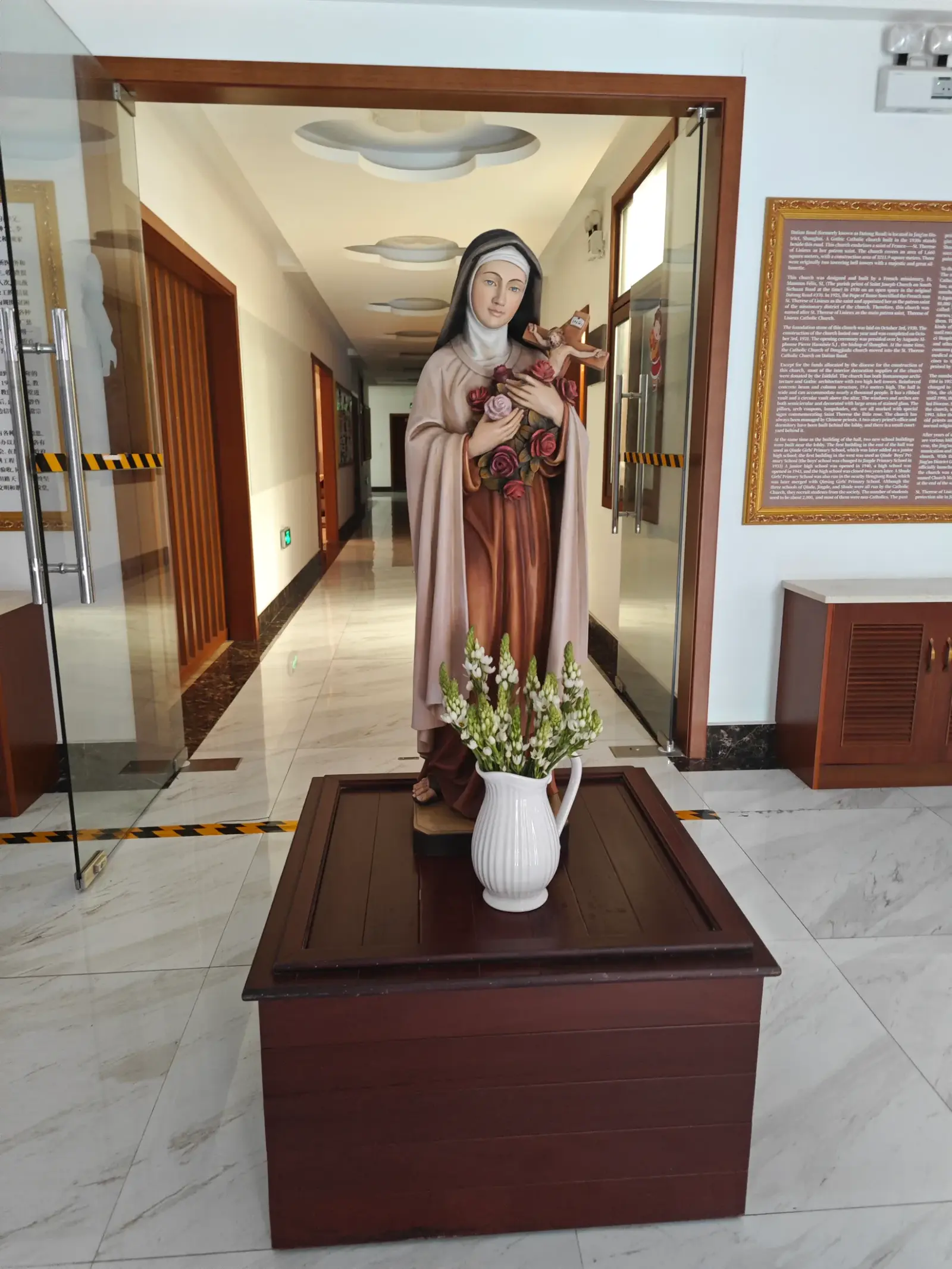 A statue of a nun holding flowers on a wooden stand in a bright hallway.