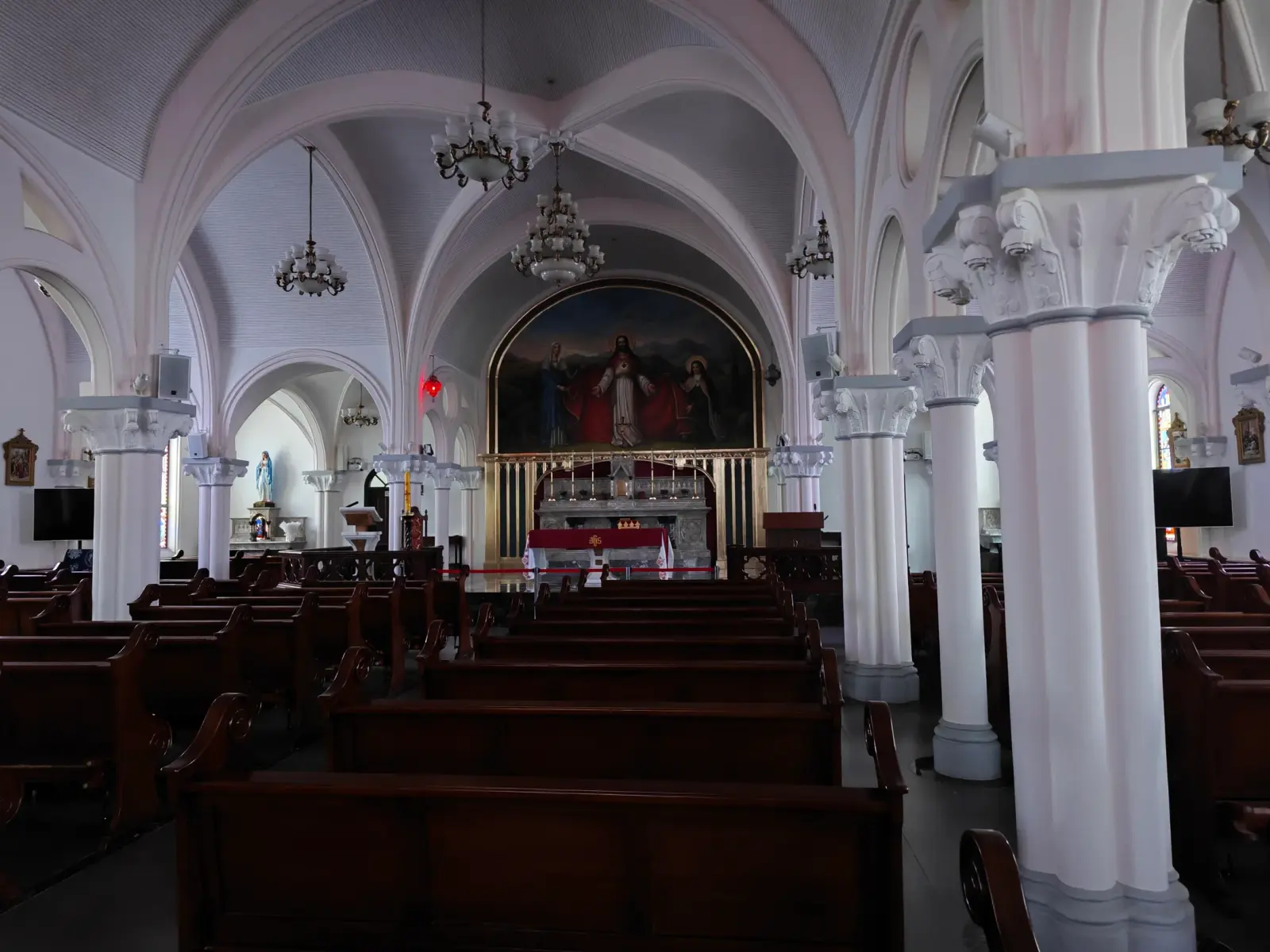 A white Gothic-style church interior with dark wooden pews and a painting above the altar.