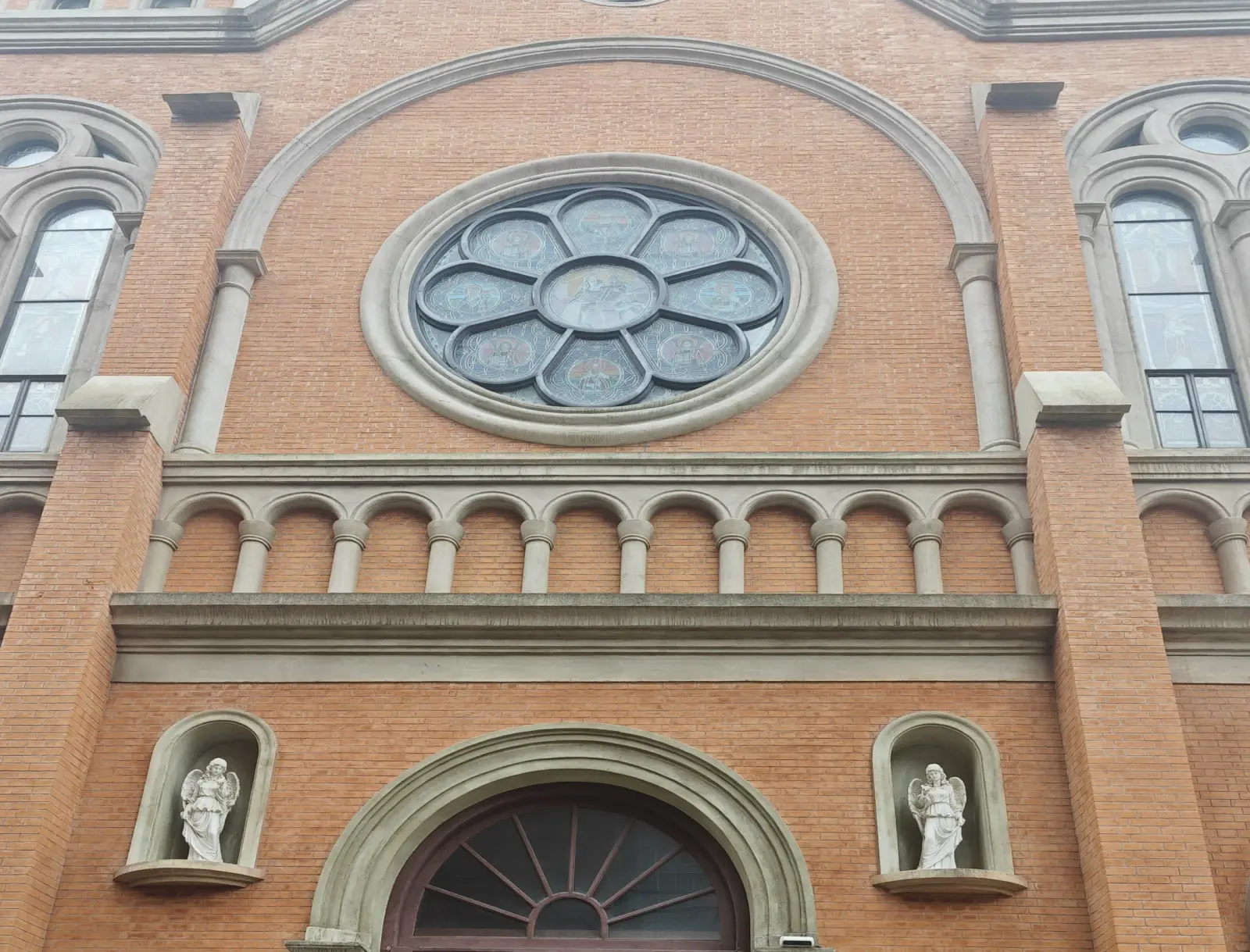Close view of an orange-brick church front with a stone rose window, a row of small arches and two white angel statues.
