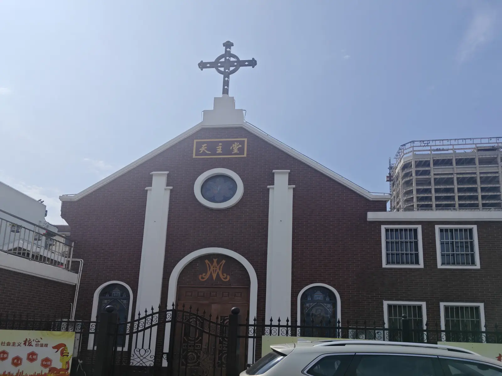 A dark red-brick church front with white pilasters, a round window, a cross on the gable and an arched wooden door behind an iron fence.