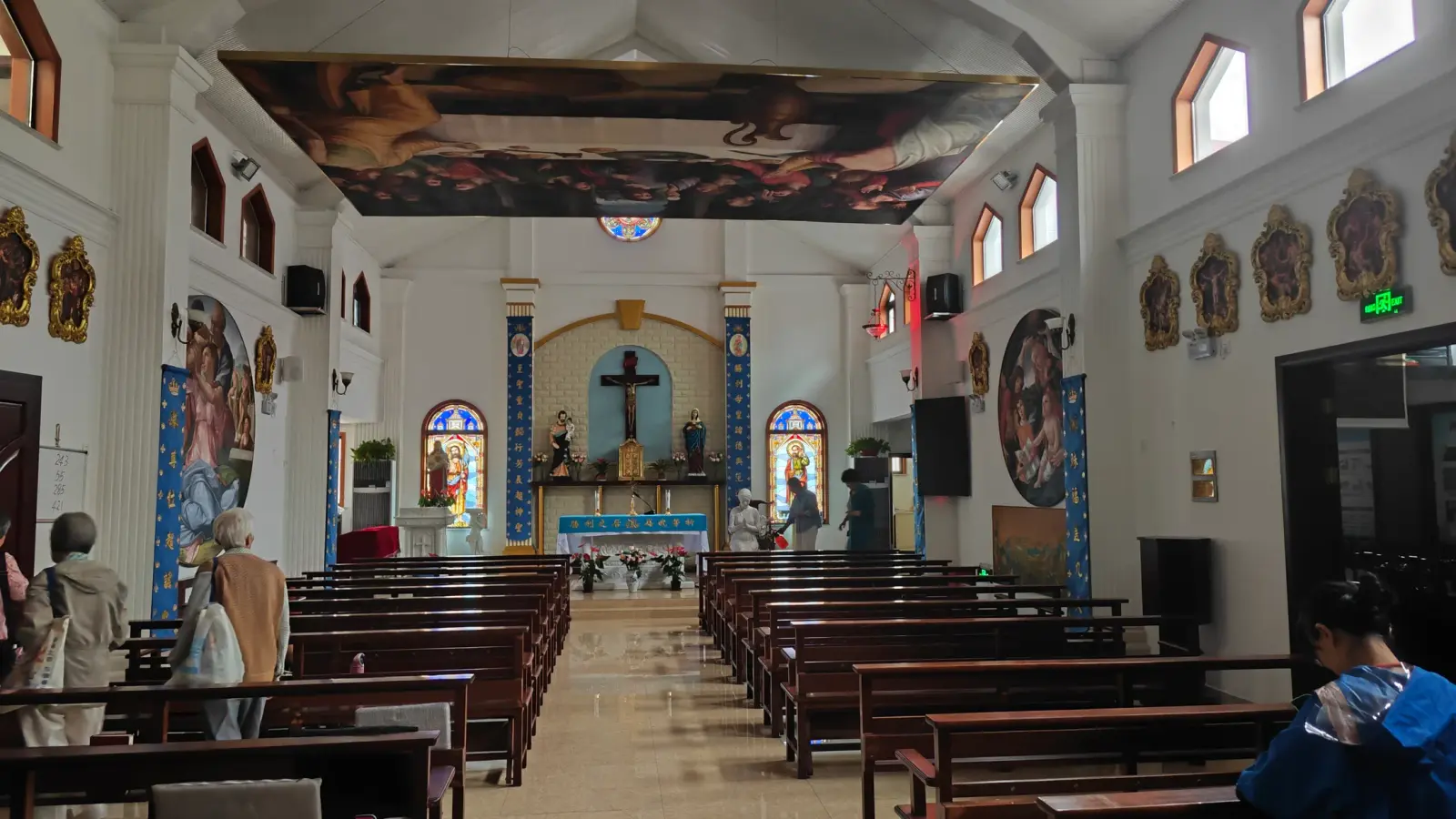 A church interior seen from the back with pews, a painted ceiling and blue-framed stained-glass windows.