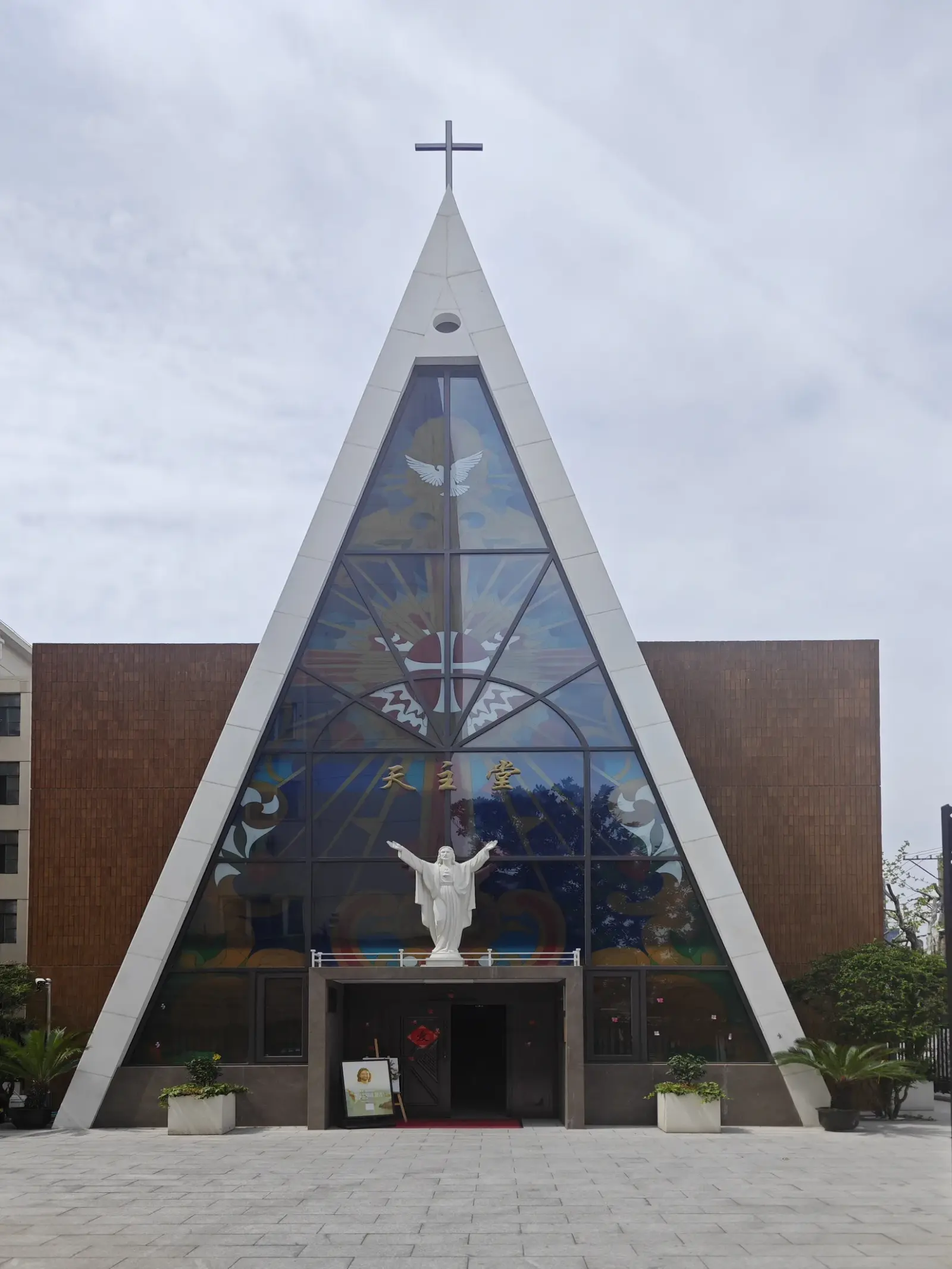 A triangular glass-and-steel church facade with stained glass, a white statue of Christ with open arms and a cross at the apex.