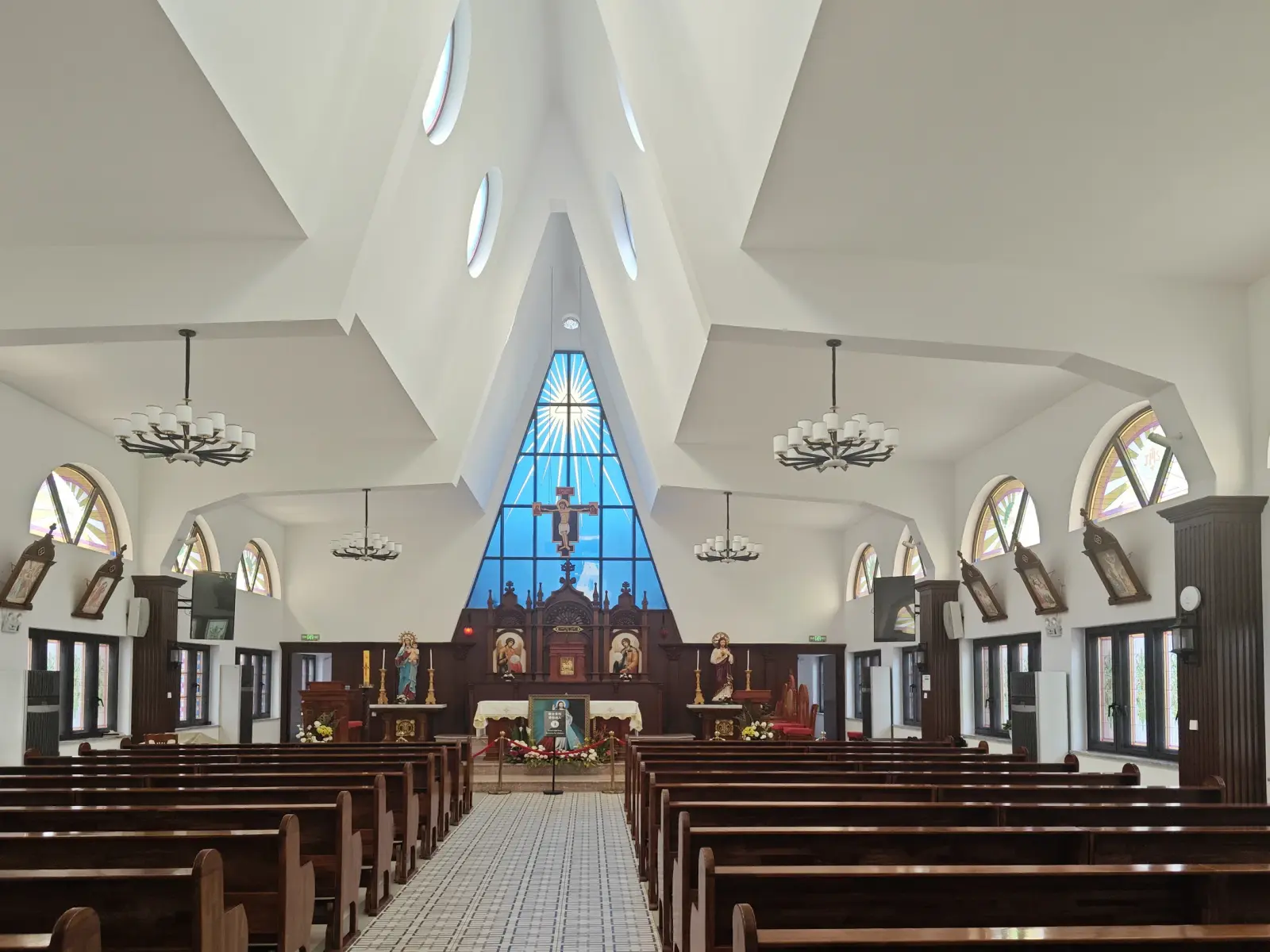 A white church interior with wooden pews, chandeliers and a blue triangular window above the altar.