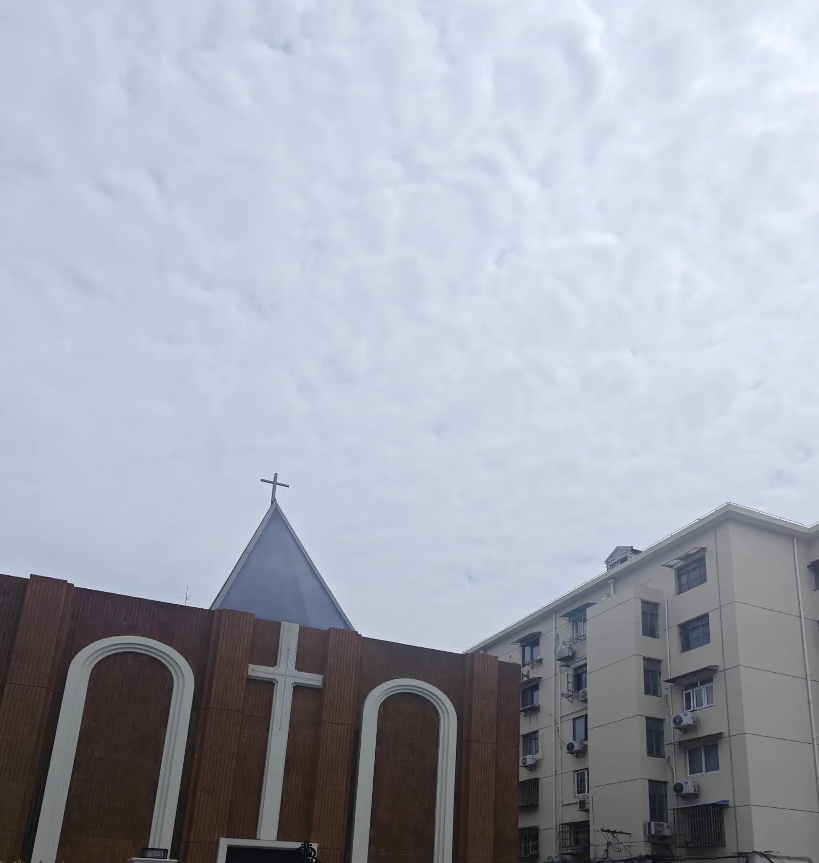 A brown wall with white arched outlines and a cross beneath a small spire, beside a block of flats.