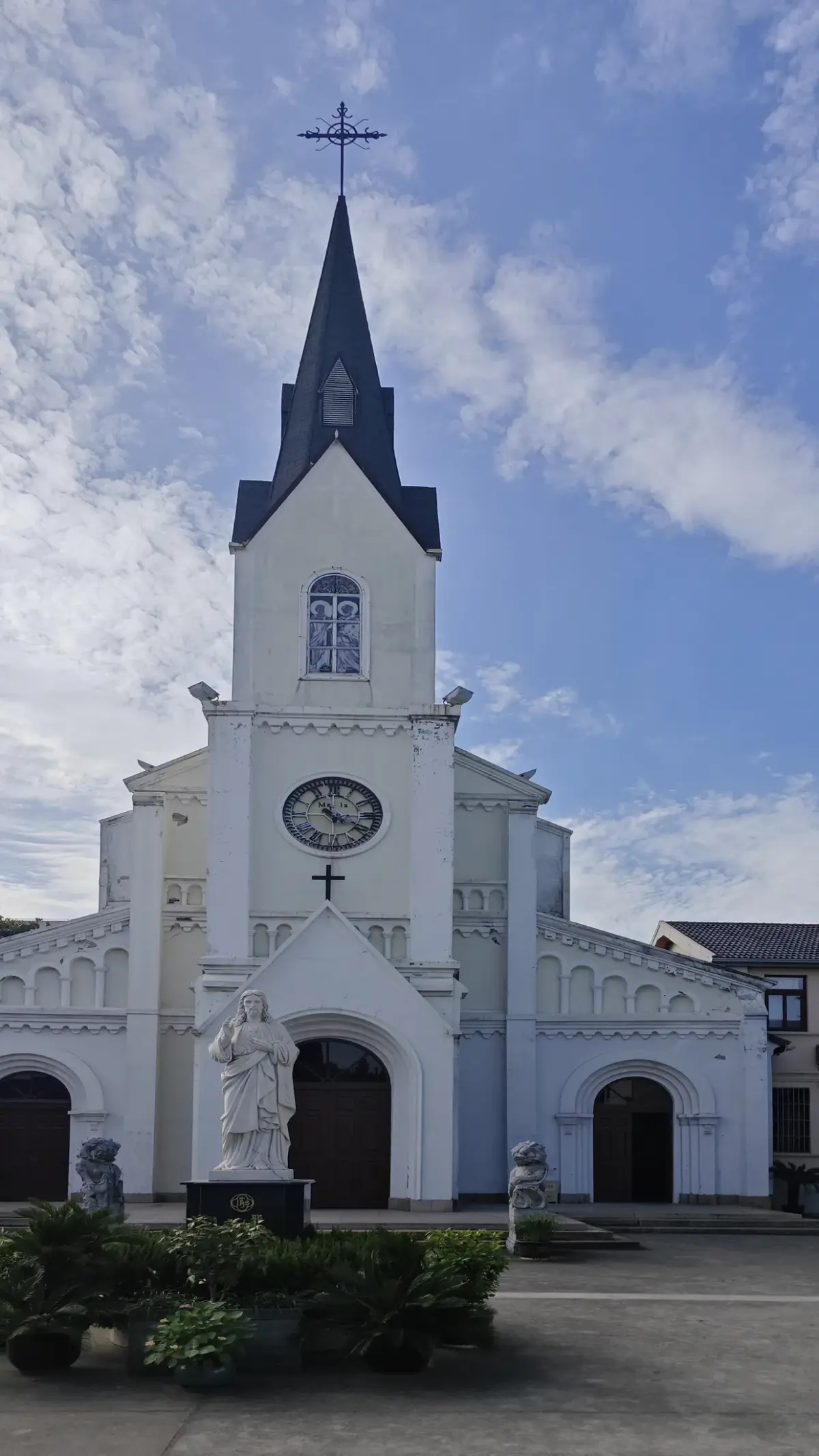 White church facade with a pointed spire, round clock and a statue of Mary in front of the entrance.