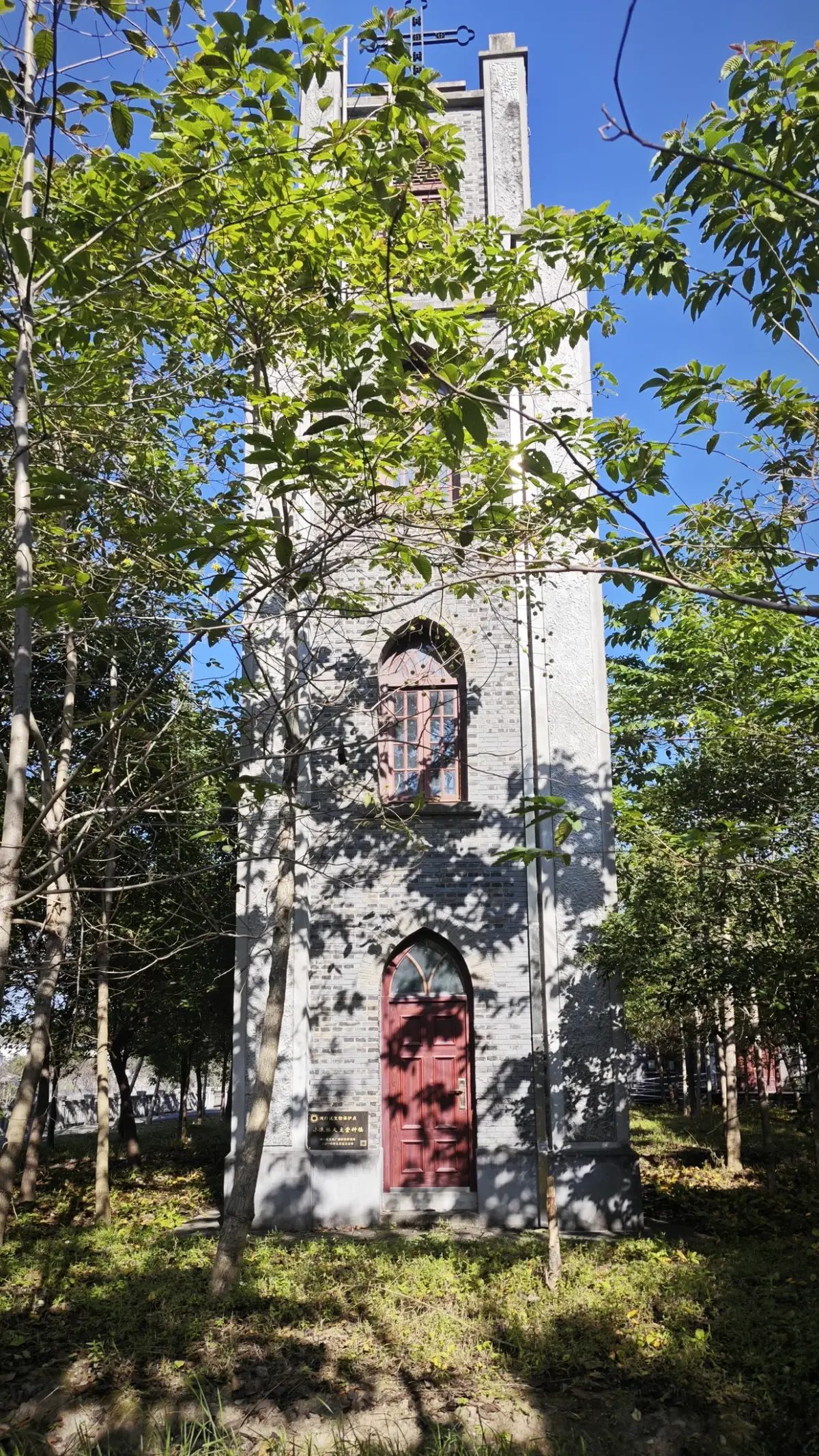 A tall narrow brick tower with pointed windows and a red door among trees.