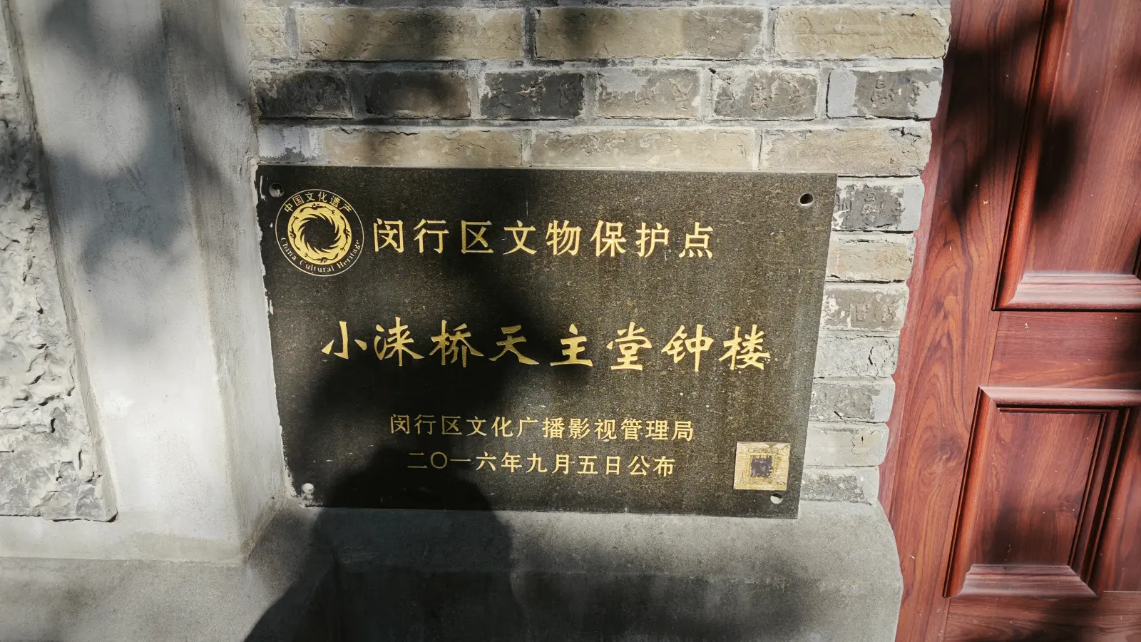 A dark plaque with gold Chinese characters on a brick wall beside a wooden door.