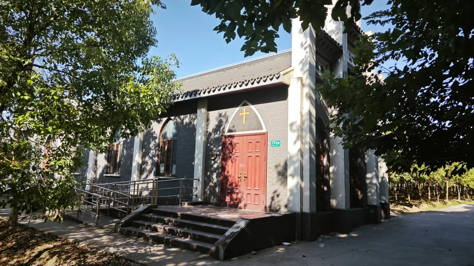 A grey church building with red double doors and steps under tree shade.