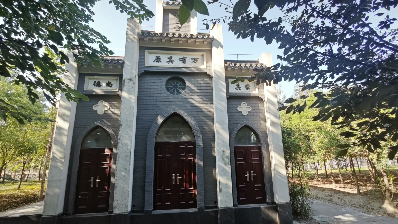 A grey-brick church front with three pointed-arch red doors, a round window and a tiled roofline, framed by trees.