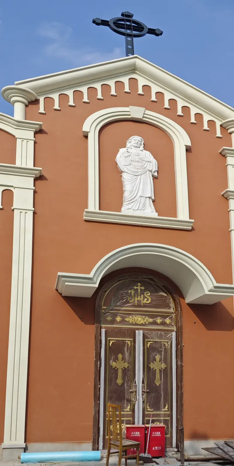 A terracotta-coloured church facade with a white statue in an arched niche, a pair of doors and a cross on top.