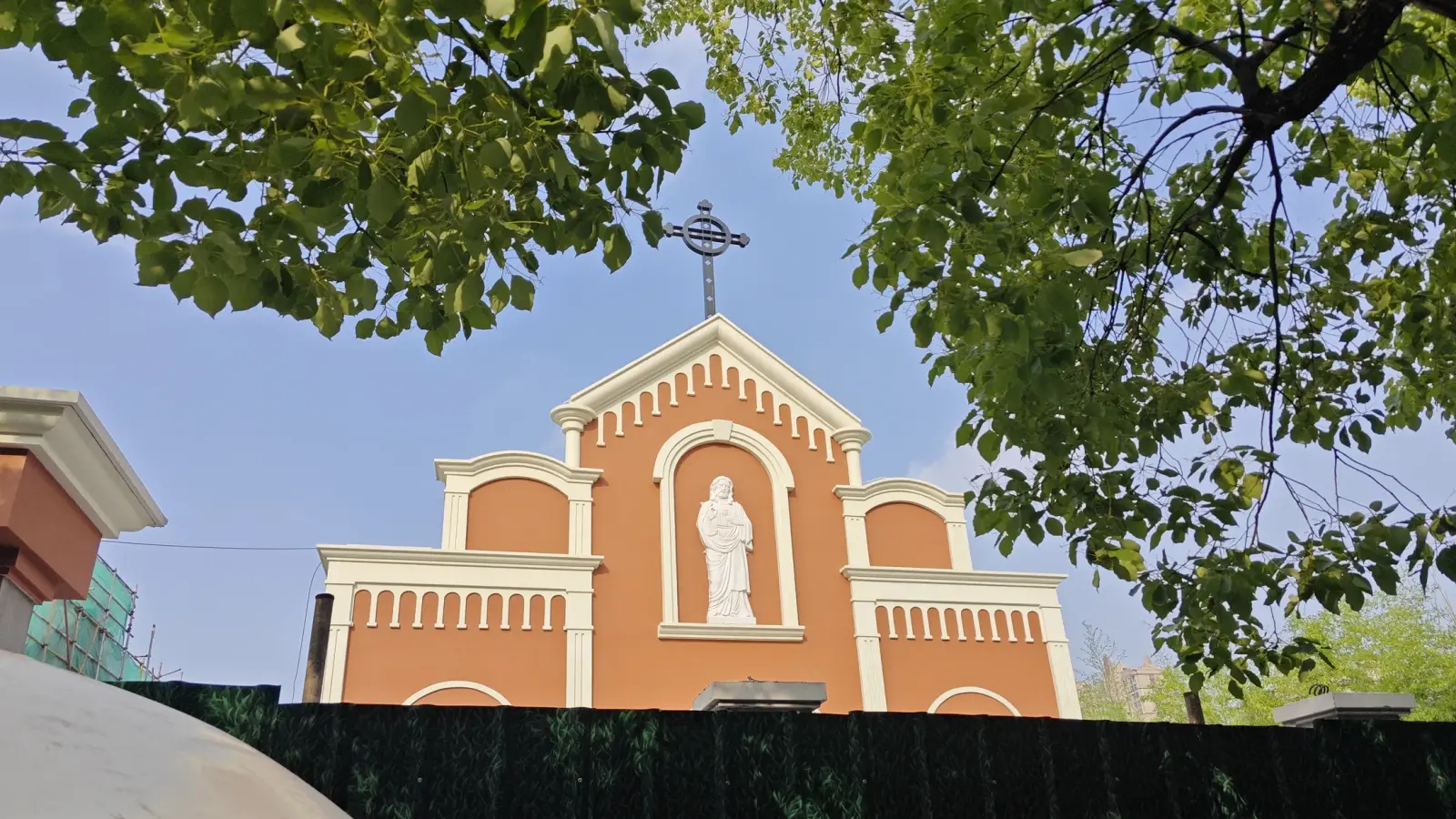 The top of an orange church front with a cross and statue seen through tree branches above a green hoarding.