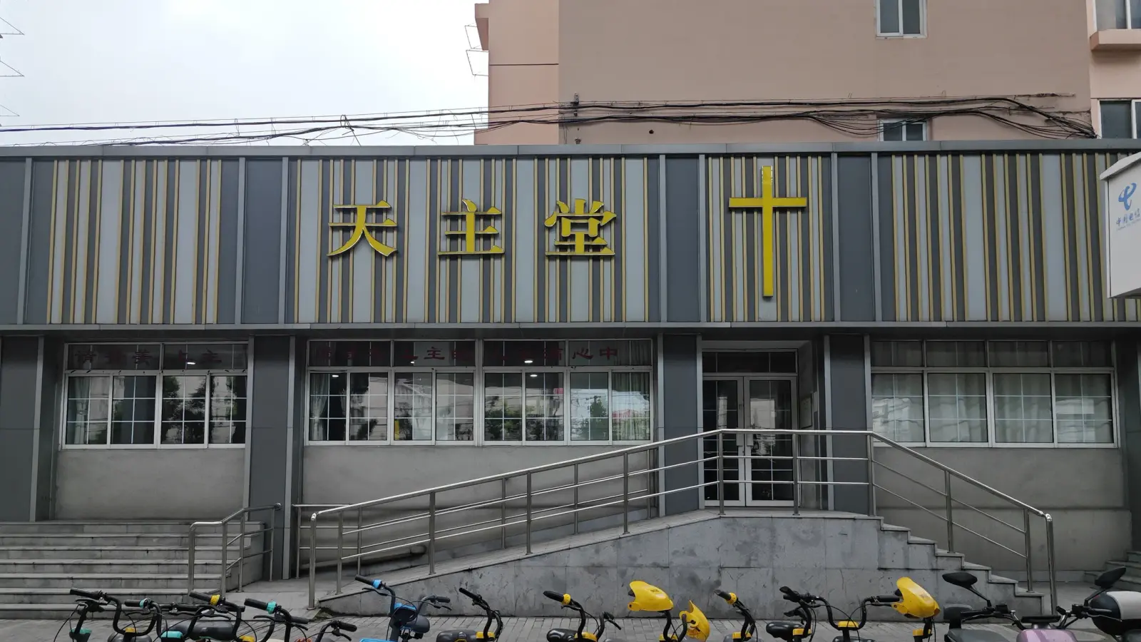 A low grey and gold striped building with a large gold cross and Chinese characters, steps and a ramp in front, and bicycles at the bottom.