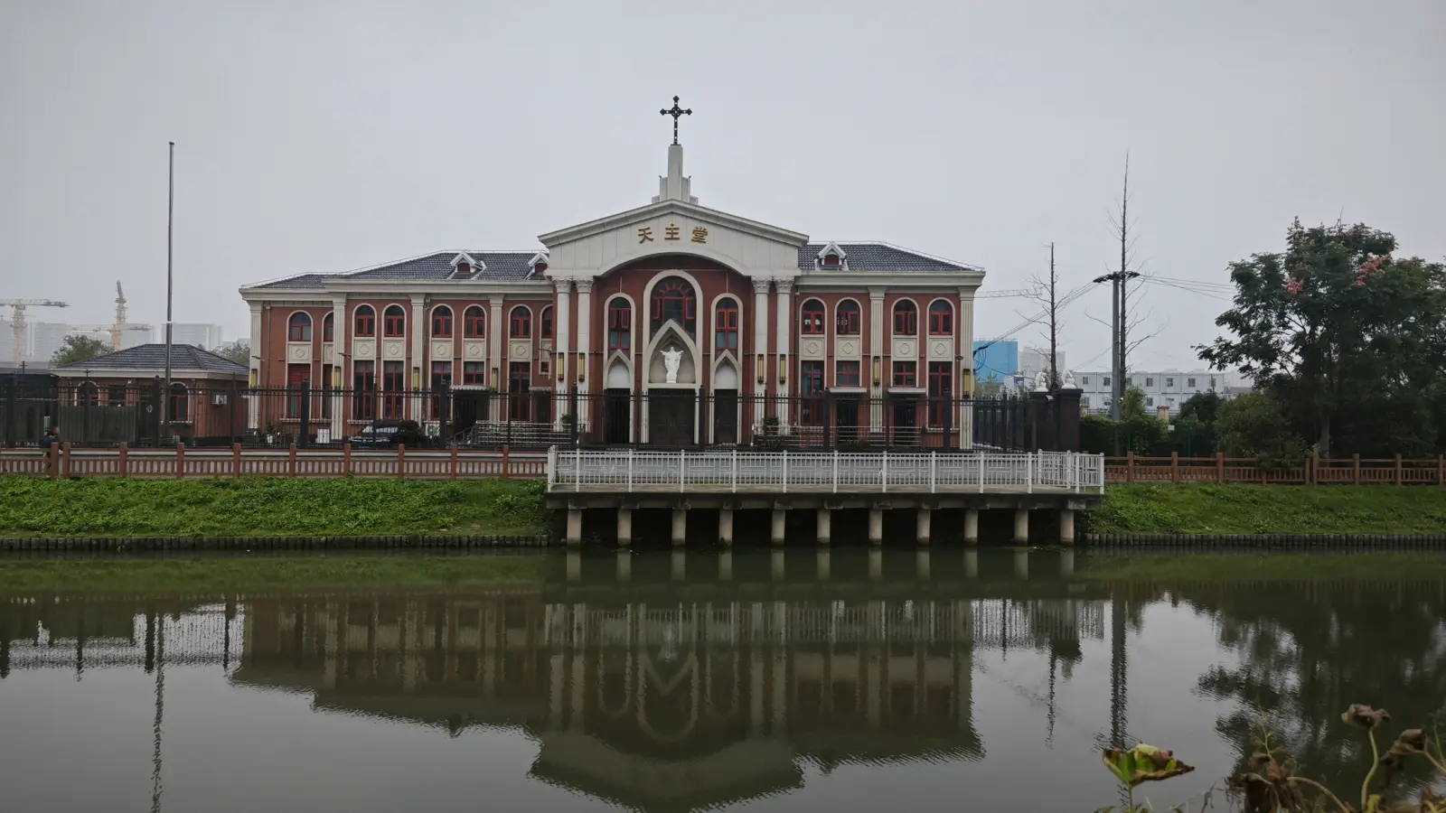A two-storey red-brick church with a white central gable and cross, seen across a canal with its reflection in the water.