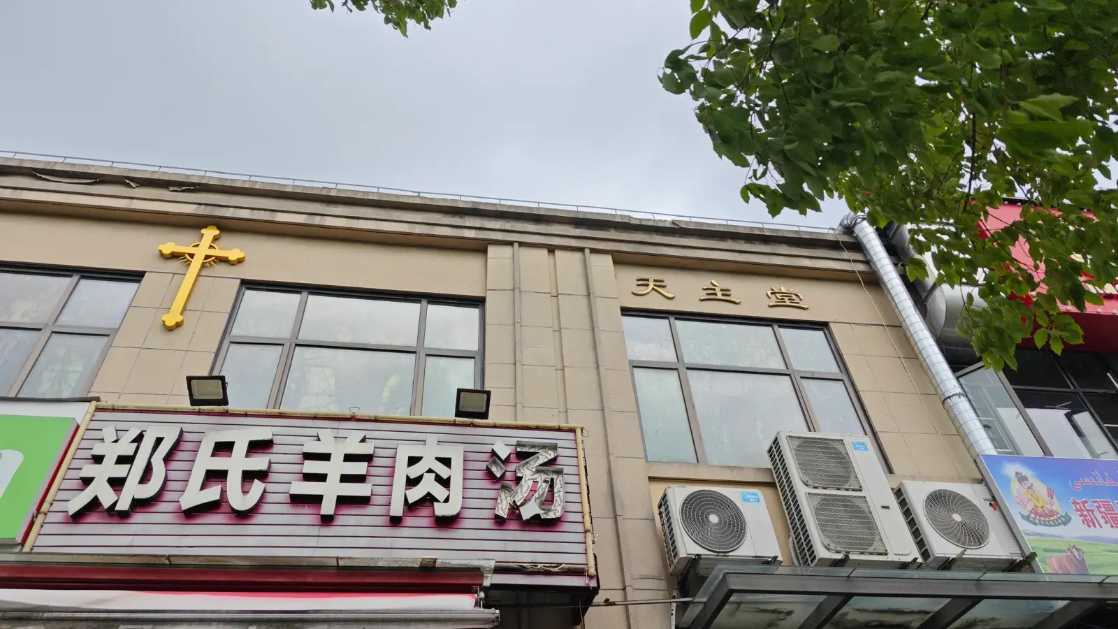 A beige two-storey building with a gold cross and characters on its upper wall above a restaurant sign.