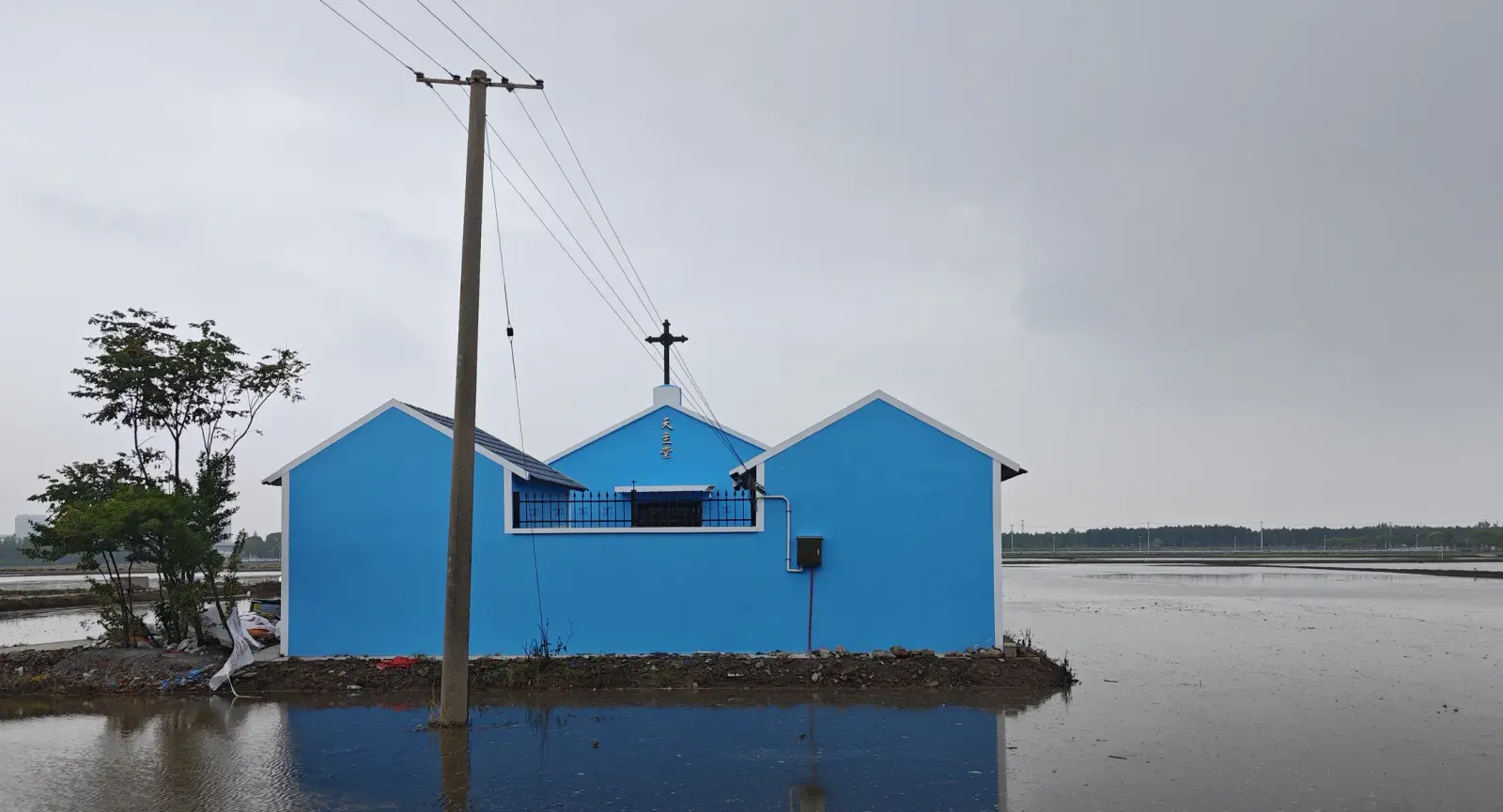 A blue church with three gables and a cross on a patch of land surrounded by water.