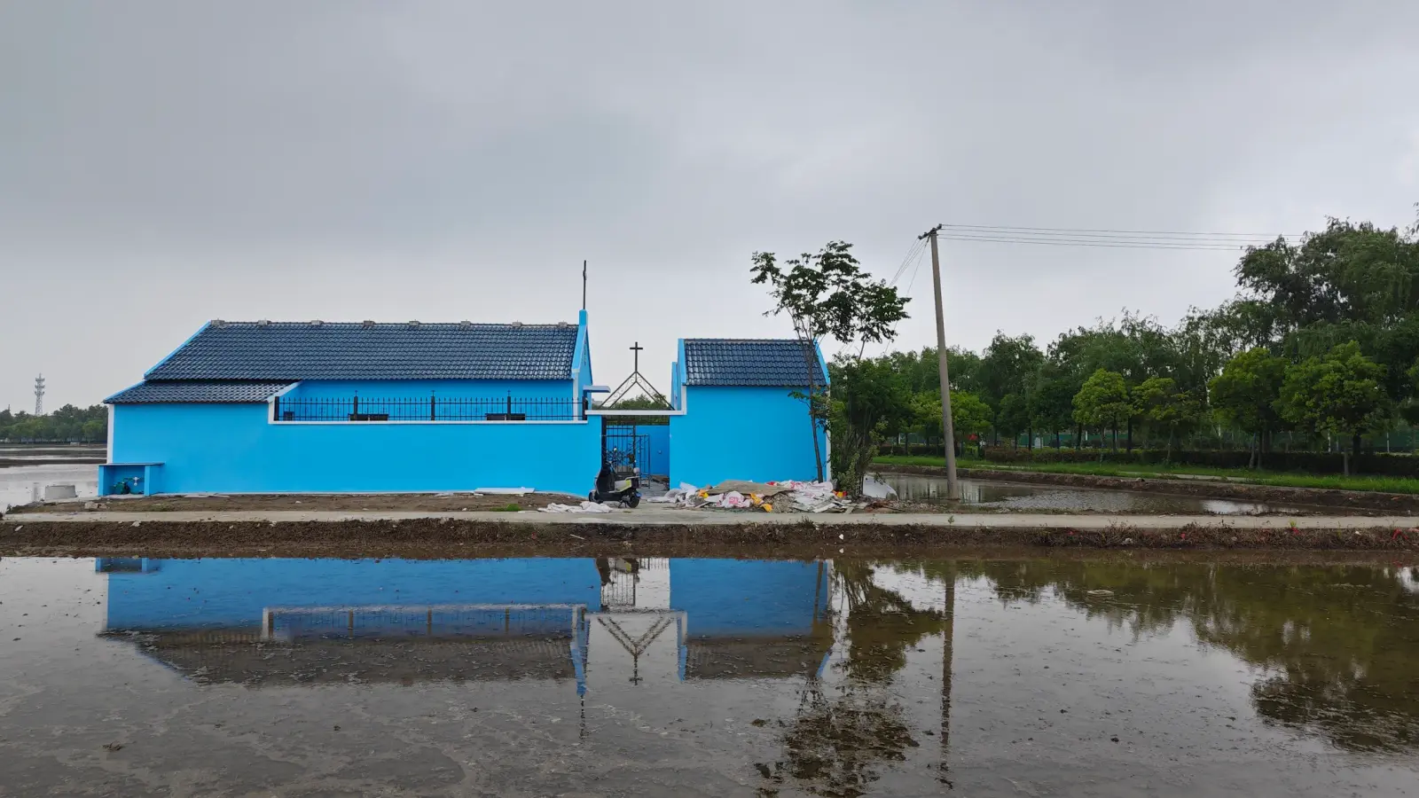 A low blue church with tiled roof and cross standing at the edge of a flooded rice field, reflected in the water.
