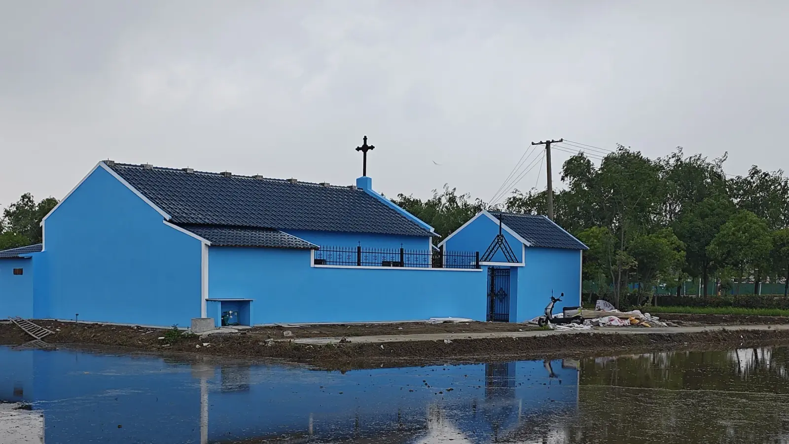 Side view of a blue church with tiled roof and cross beside a flooded field.