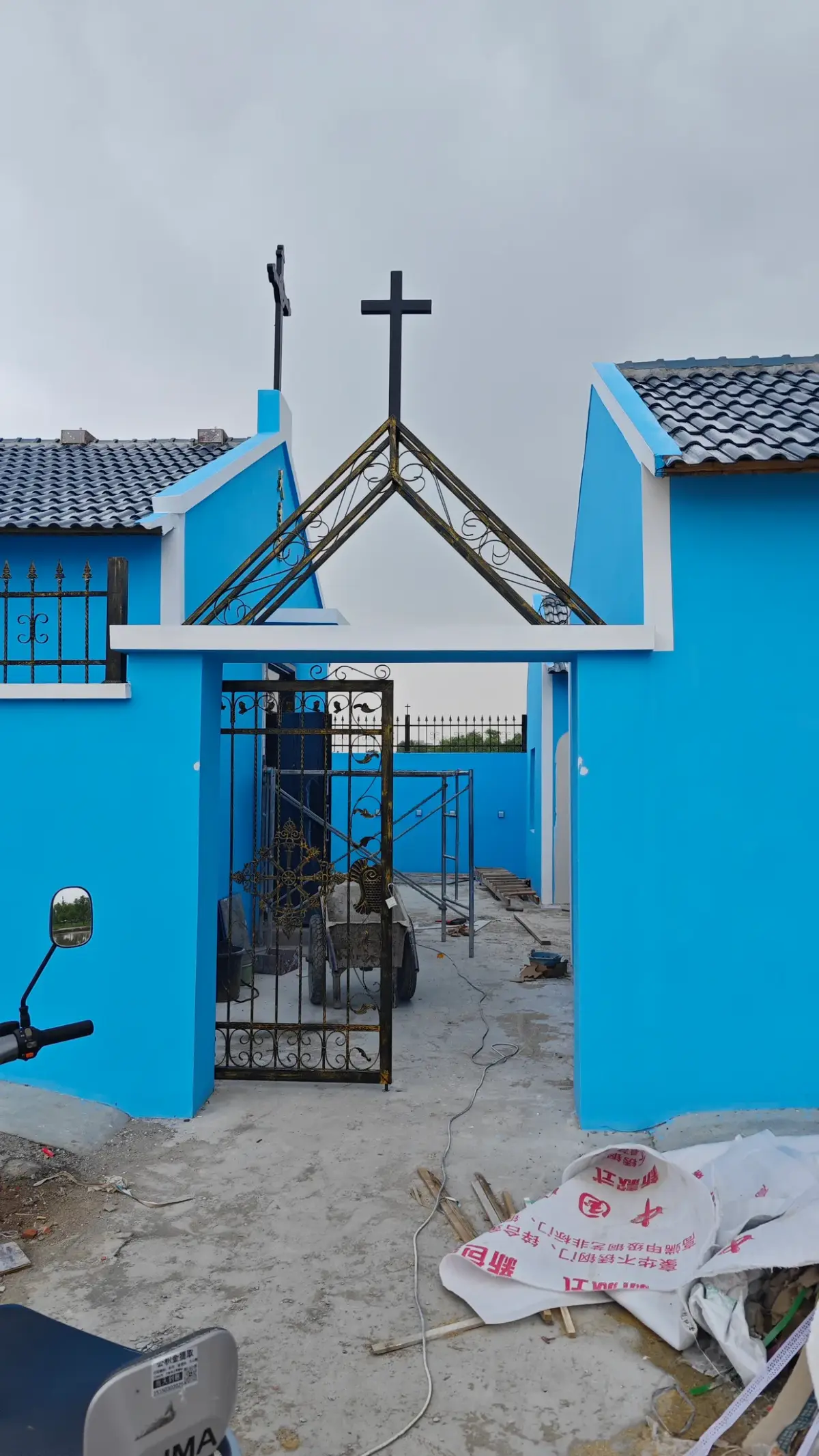 A gateway with a pointed iron frame and crosses on the roofline of a blue church.