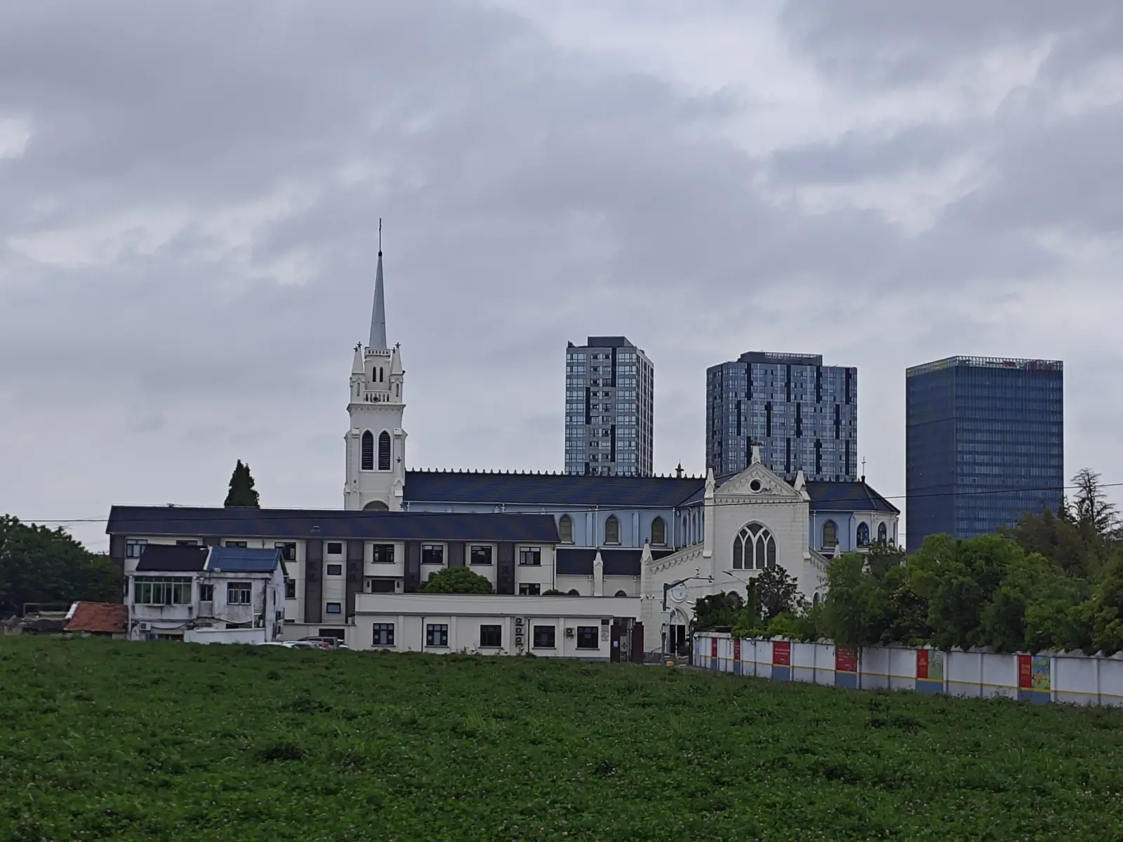 A tall white church spire and long grey-roofed buildings behind a green field, with modern office towers beyond.
