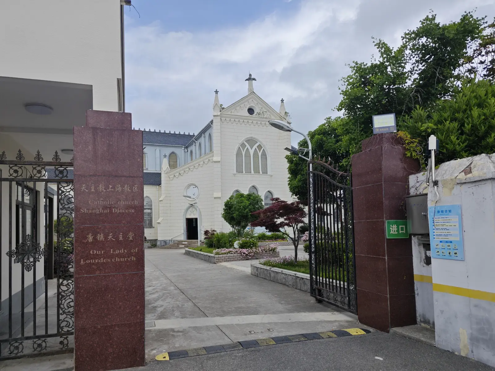 A polished red granite gate post with engraved Chinese and English lettering beside an iron gate, and a white Gothic church beyond.
