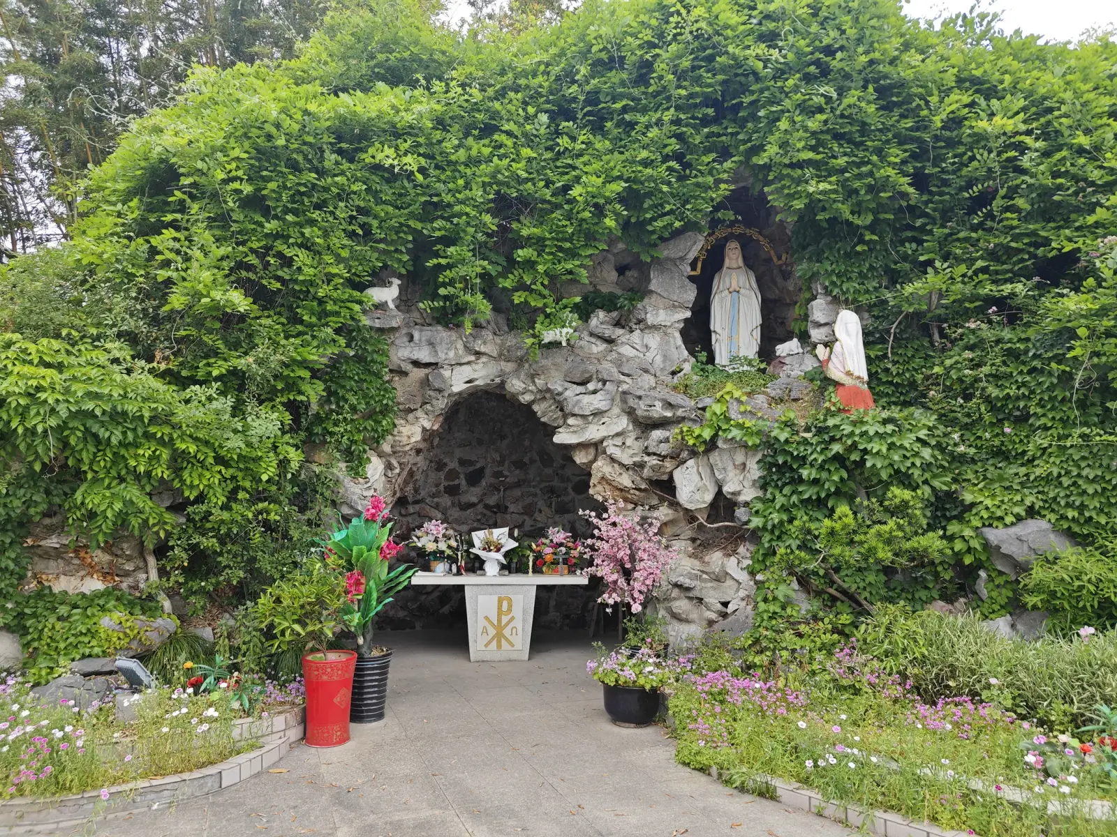 A rock grotto overgrown with green vines, with a white statue in a niche and an altar with flowers.