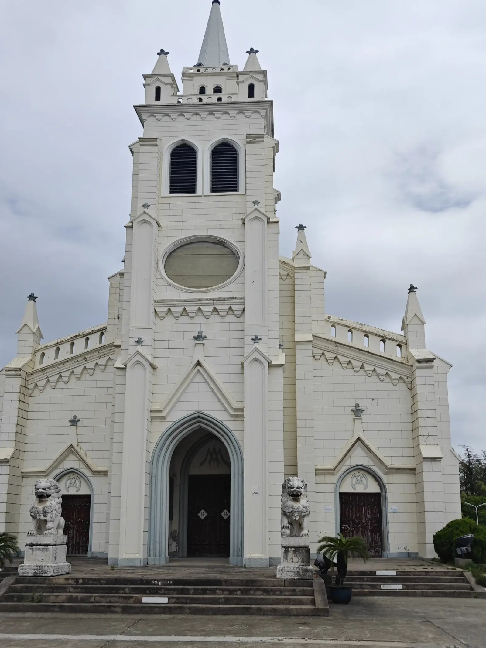 A tall cream church tower with a spire above a pointed-arch entrance flanked by stone lion statues and entrance steps.