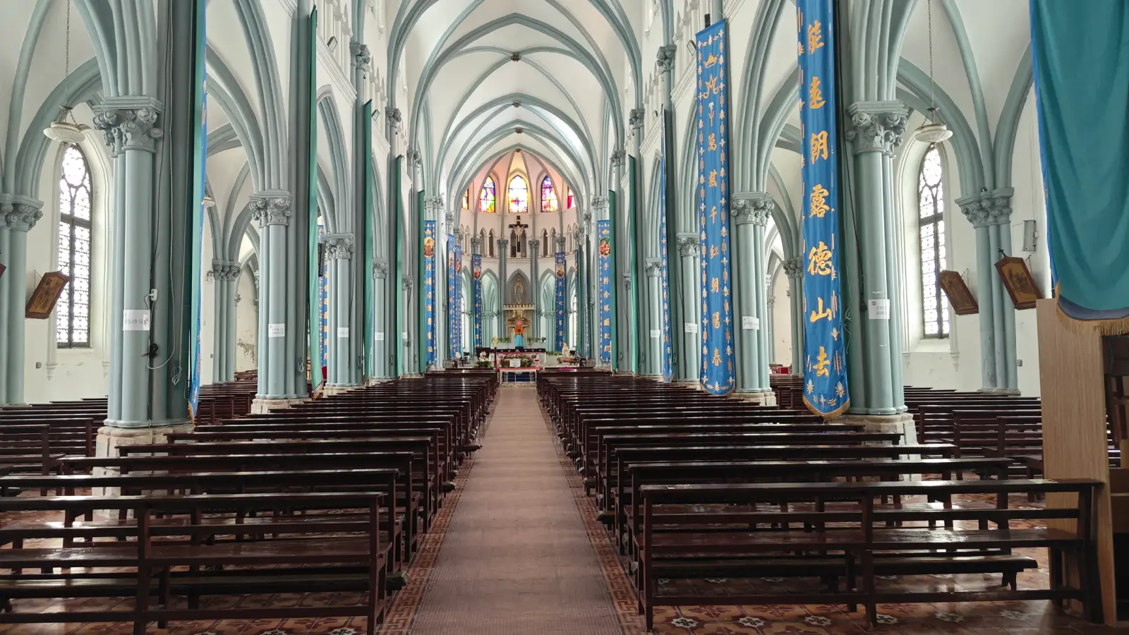 A long Gothic church interior with pale green vaulting, rows of dark pews and a lit stained-glass window at the far end.