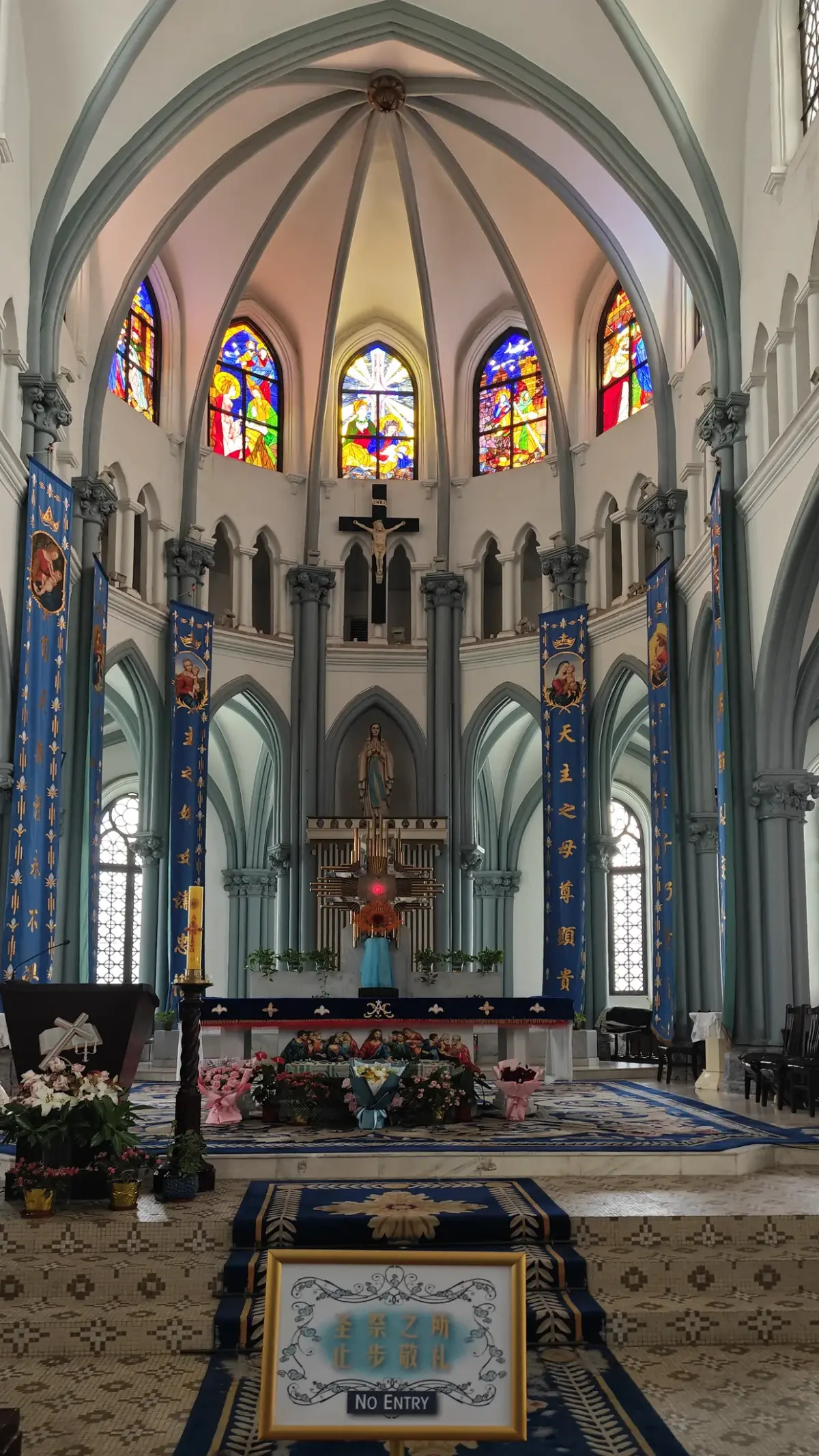 A Gothic apse with ribbed vaulting, small stained glass windows, blue banners and an altar on a patterned carpet.