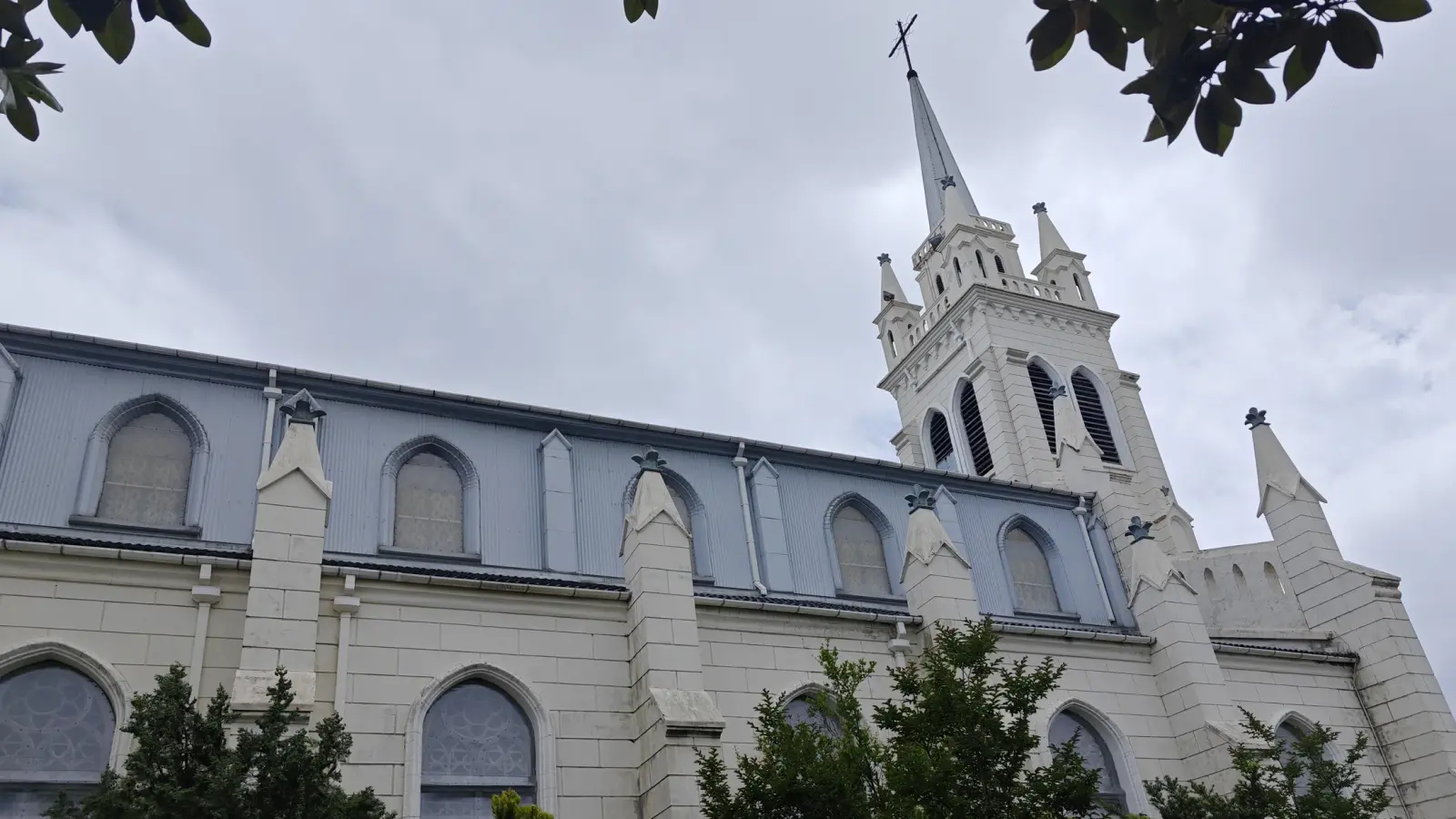 Looking up at a cream Gothic church tower and spire above a wall of pointed windows and buttresses.