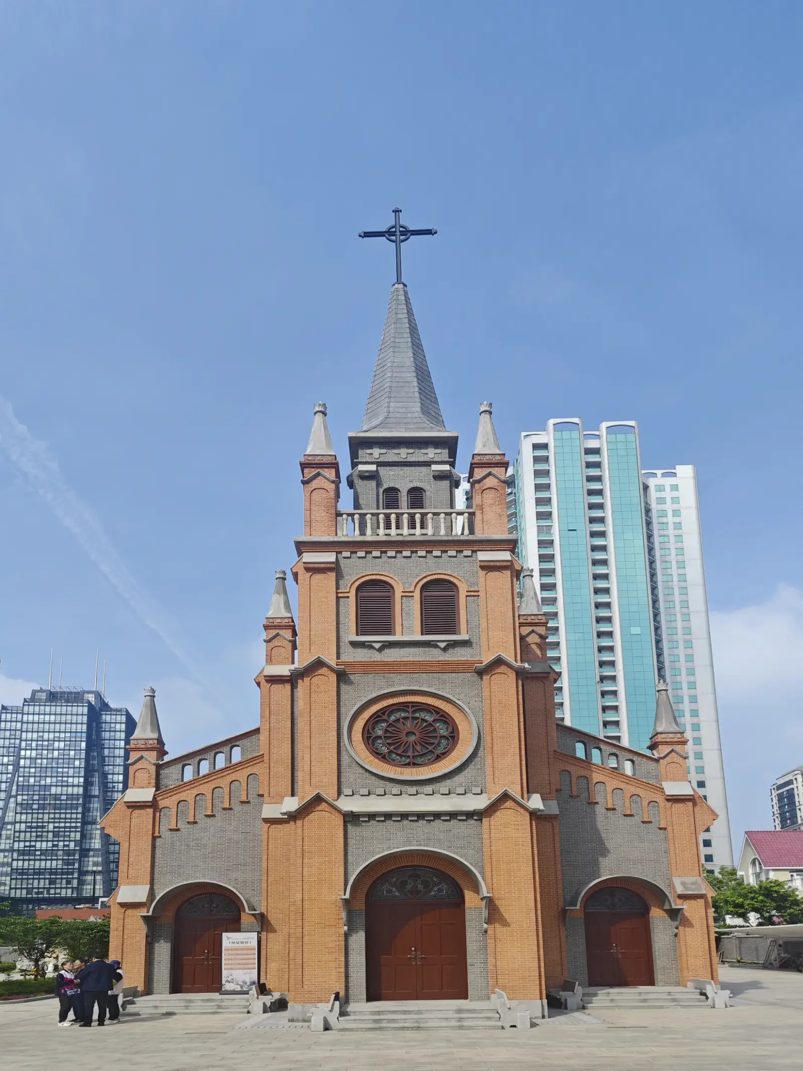 A red-orange brick church front with grey brick bands, a central tower with a pointed spire and a rose window above two arched doorways.