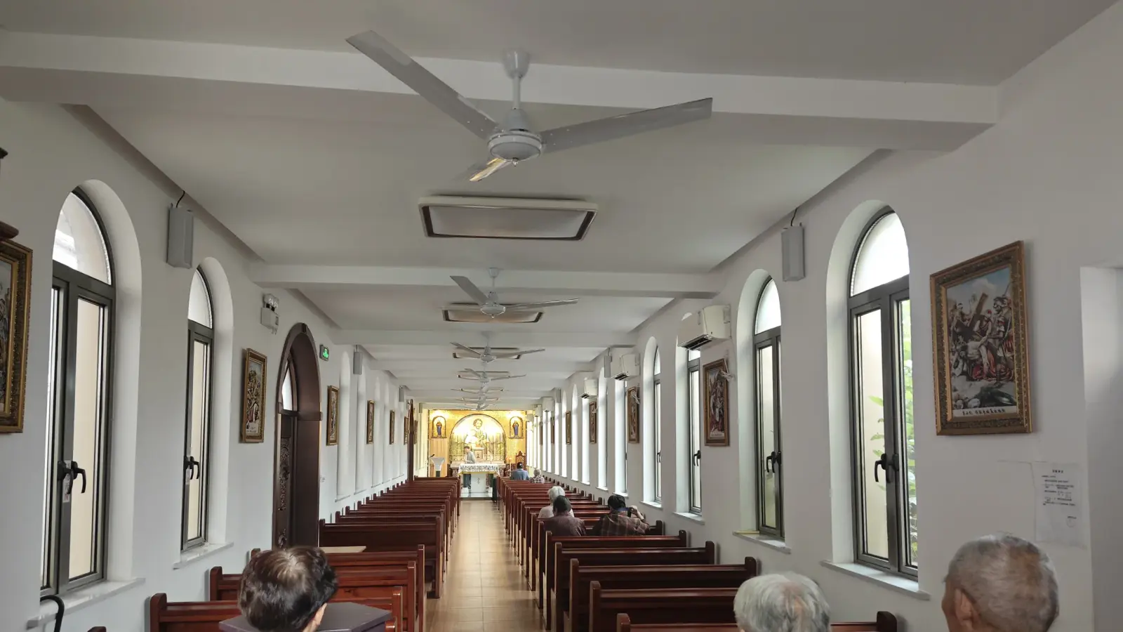 A narrow church interior with tall arched windows, framed pictures on the walls, wooden pews and an altar glowing gold at the far end.