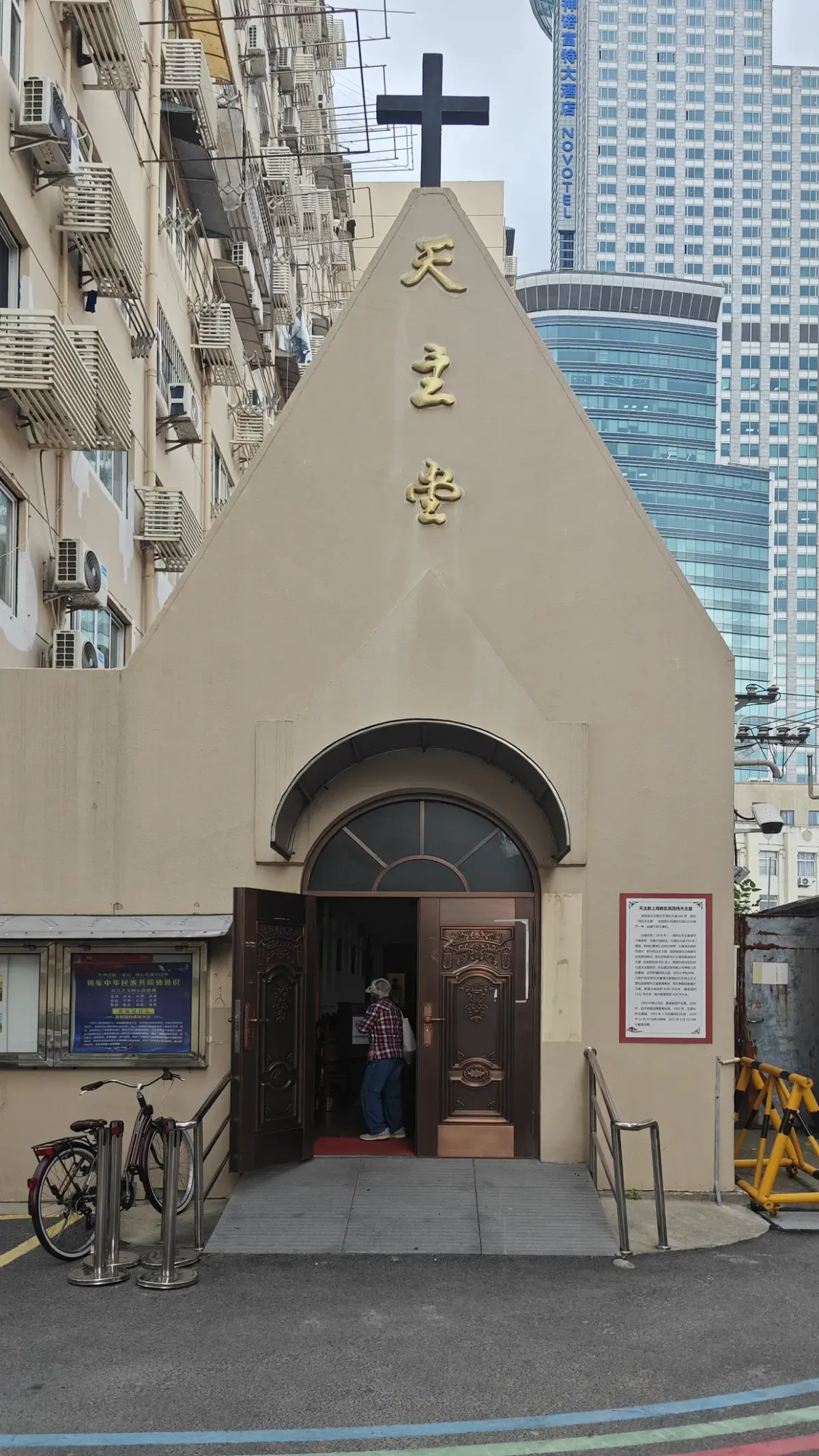 A small church front with a cross, an open brown door and notice boards, in a lane painted with rainbow stripes.