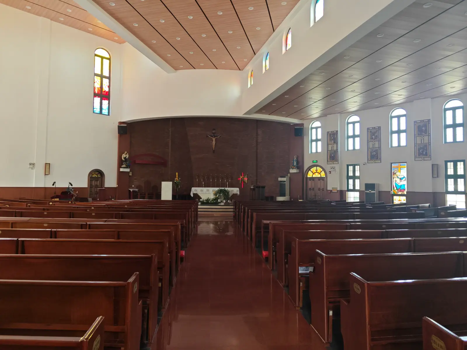 A bright church interior with long wooden pews, arched windows with coloured glass and a brown wall behind a crucifix.